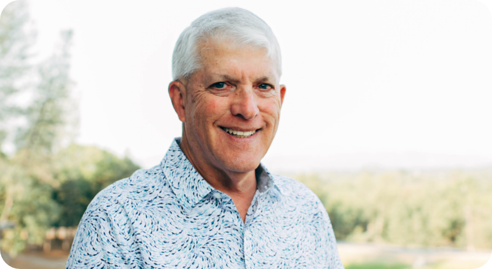 A smiling middle-aged man with white hair, wearing a light blue patterned shirt, standing outdoors with a blurred natural background.