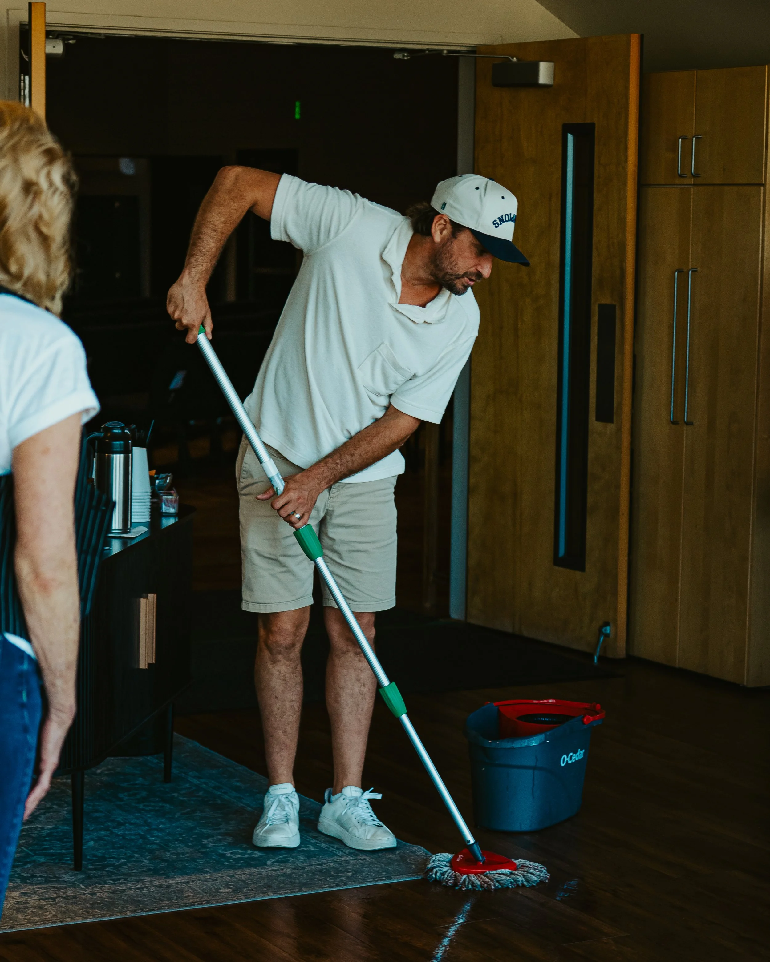 A man wearing a white baseball cap, white polo shirt, beige shorts, and white sneakers mops a wooden floor with a red and gray mop. A woman with curly blonde hair, wearing a white shirt, stands nearby. The room has wooden cabinets, a black trash bin, and a black cabinet with a small kettle and cups, indicating a kitchen or break room.