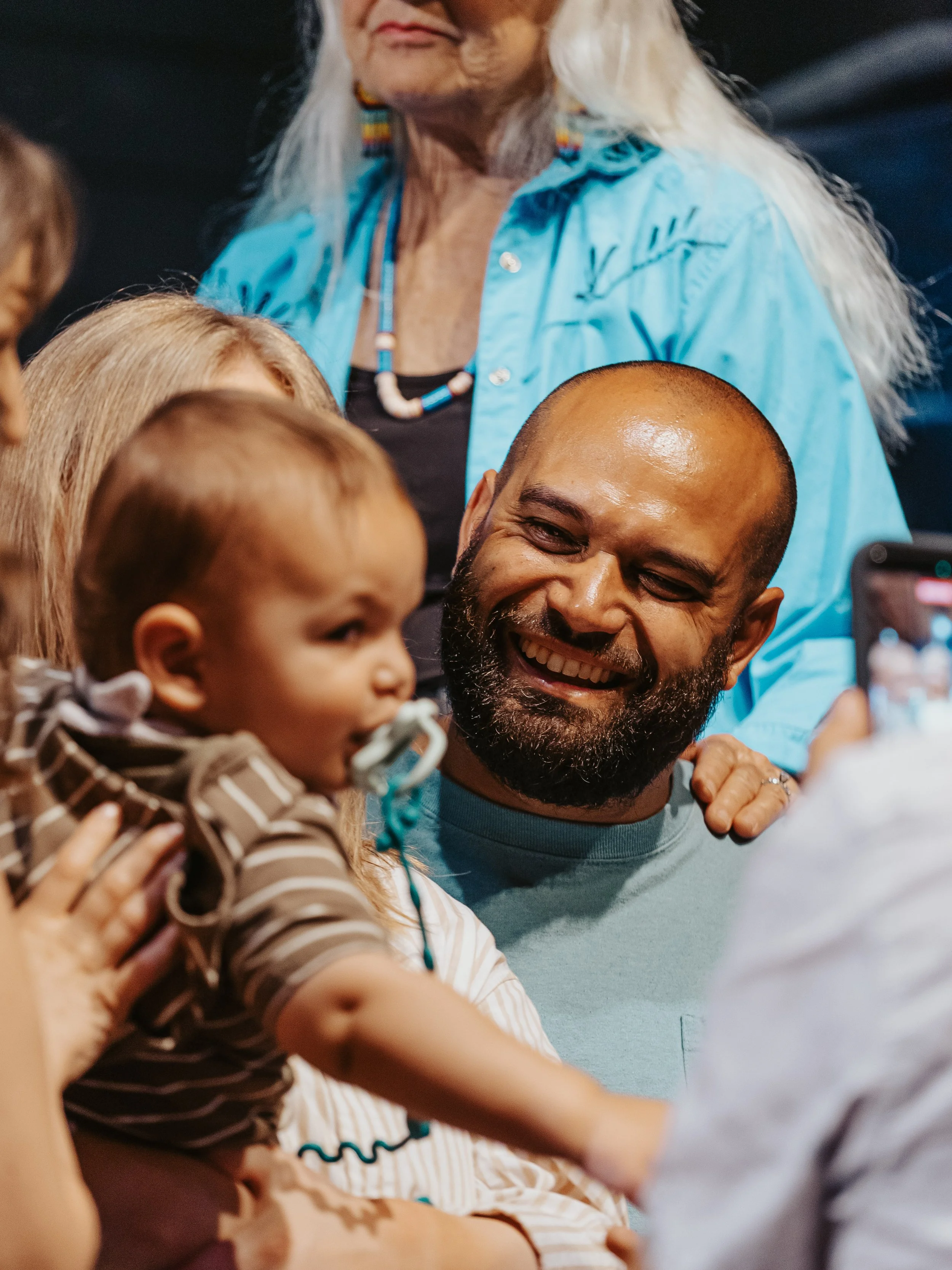 A group of people sharing a joyful moment, with a focus on a smiling bald man with a beard, holding a child with blonde hair. The background shows a woman with long white hair wearing a blue shirt and colorful earrings.
