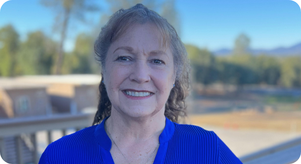 A woman with curly red hair smiling outdoors on a sunny day, wearing a blue top, with trees and a building in the background.