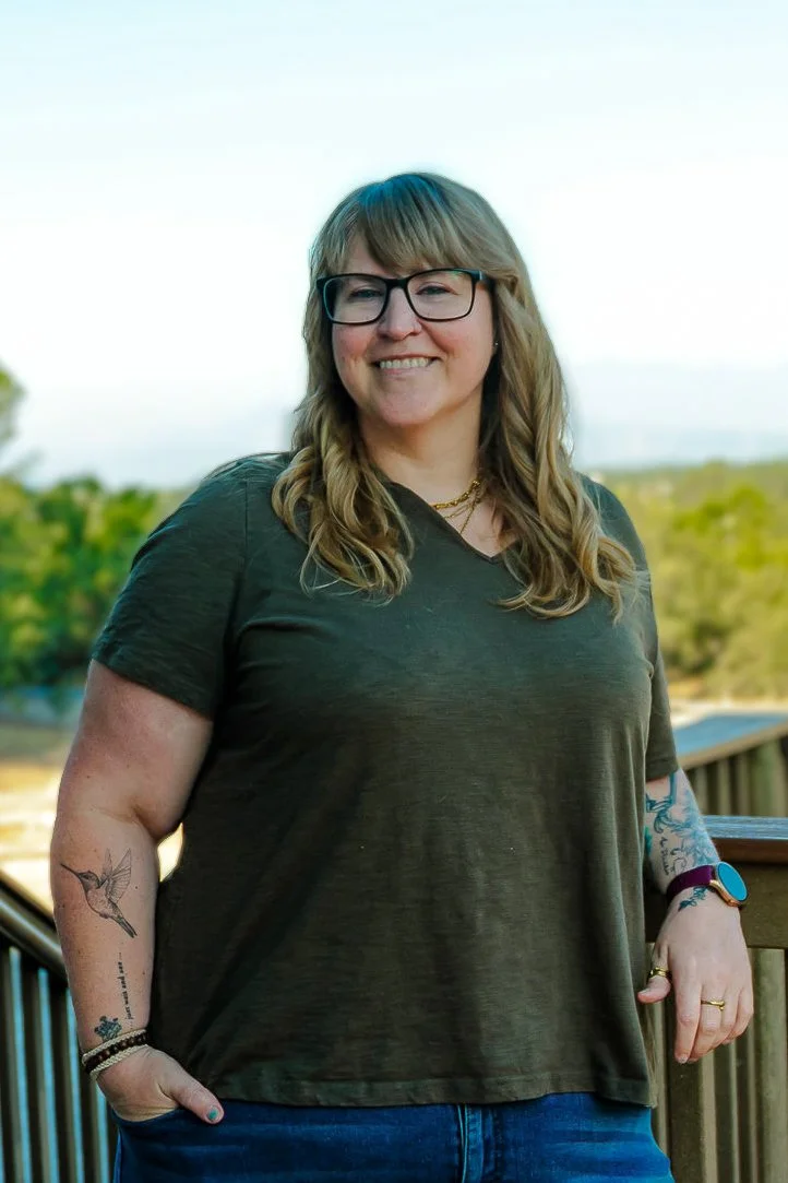 A woman with glasses, long wavy hair, and tattoos on her arms, smiling outdoors on a wooden deck with greenery in the background.