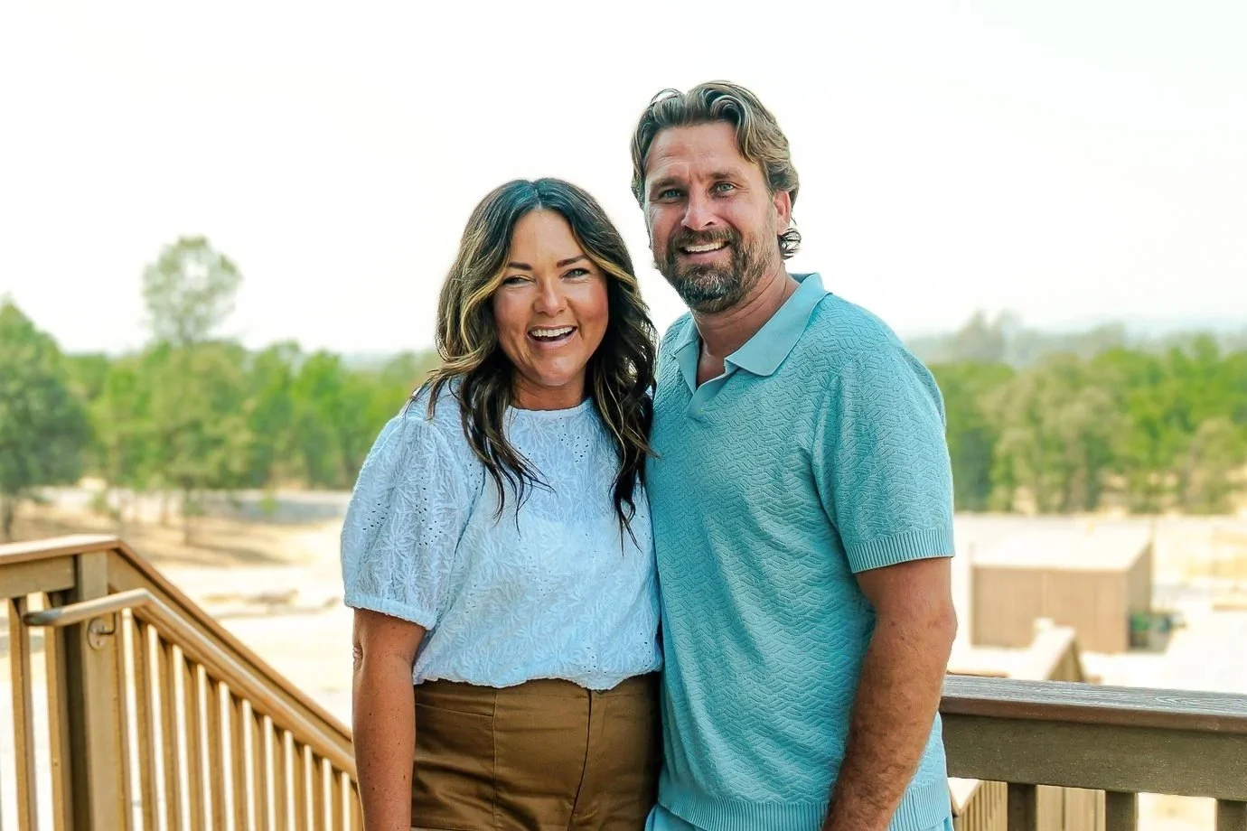 A smiling woman and man standing outdoors on a wooden deck with blurred trees and sky in the background.
