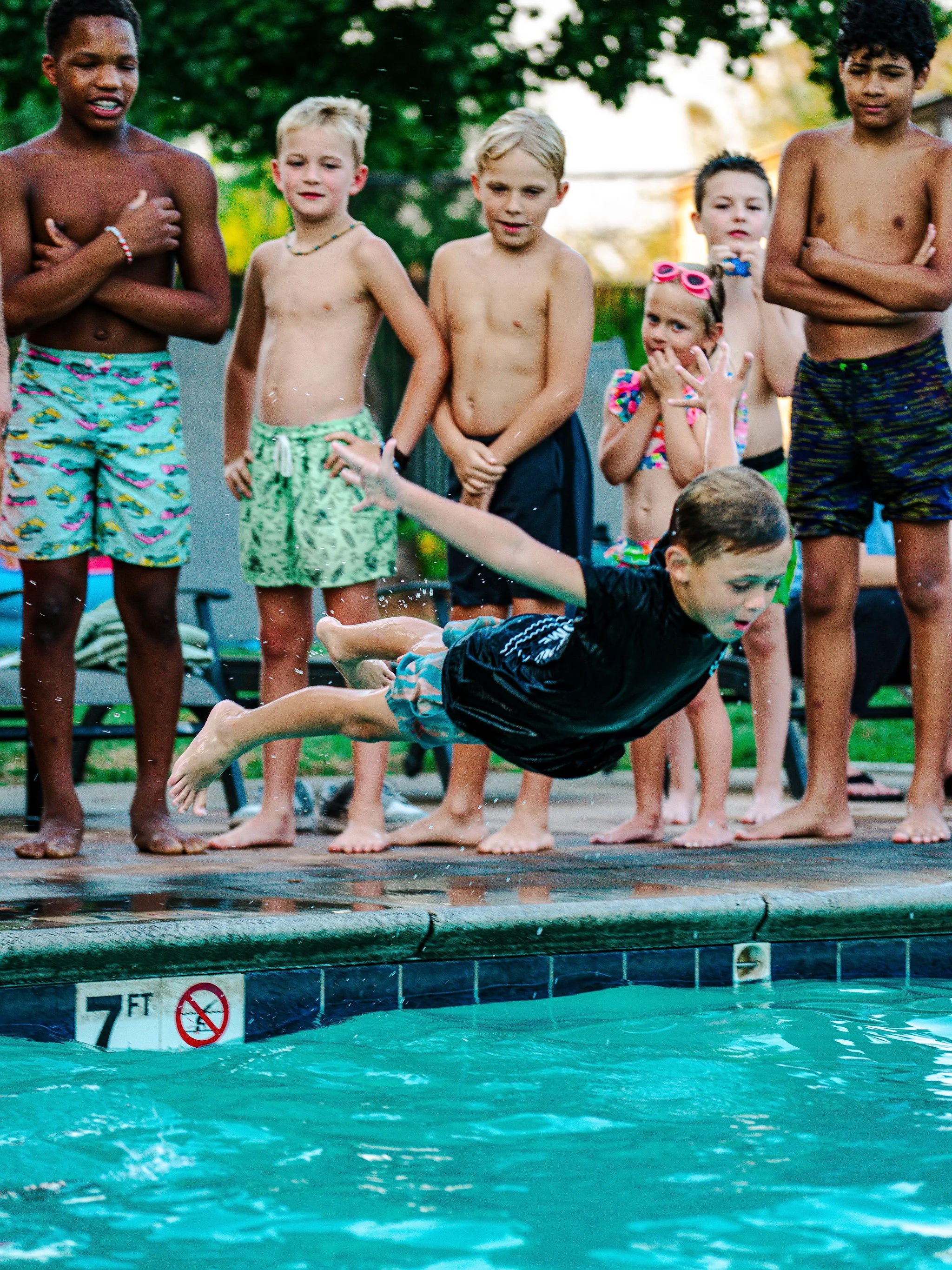 Group of children in swimsuits standing at the edge of a swimming pool, watching a boy dive into the water.
