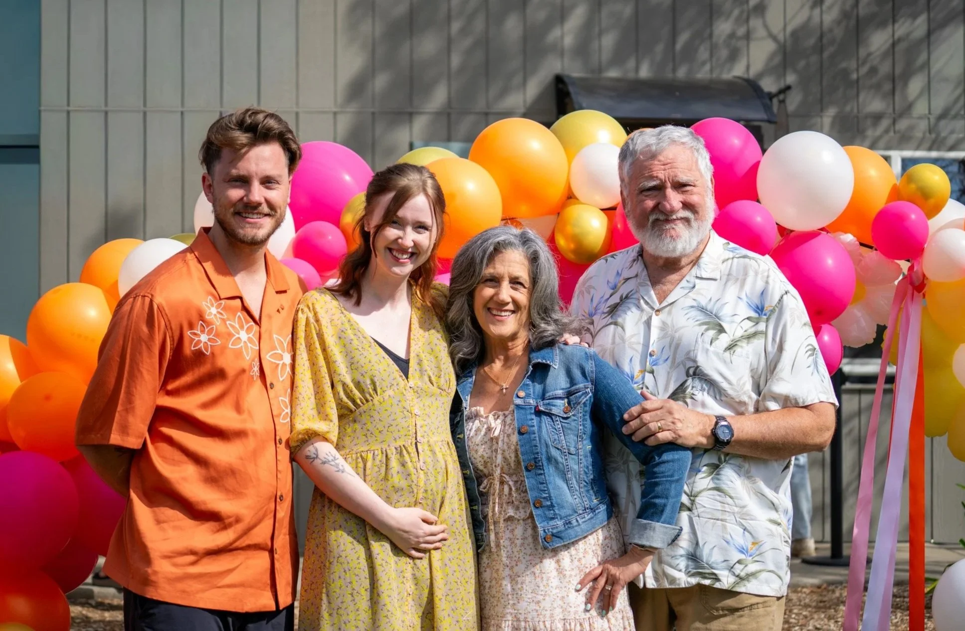 Group of five smiling people standing in front of a colorful balloon display, outdoors in daytime.
