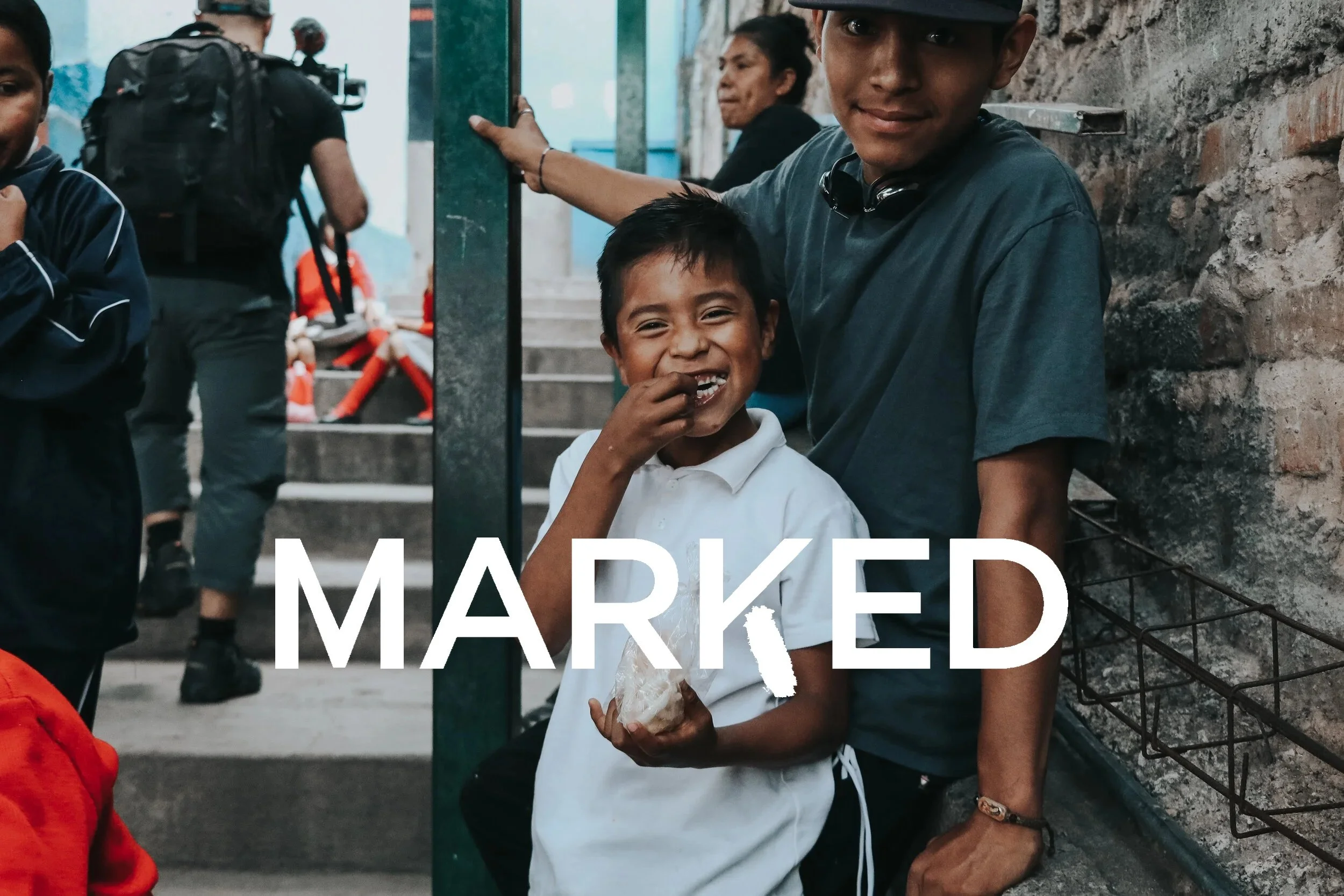 Two boys smiling and posing for the camera outdoors, one holding a piece of bread, with a group of people in the background.