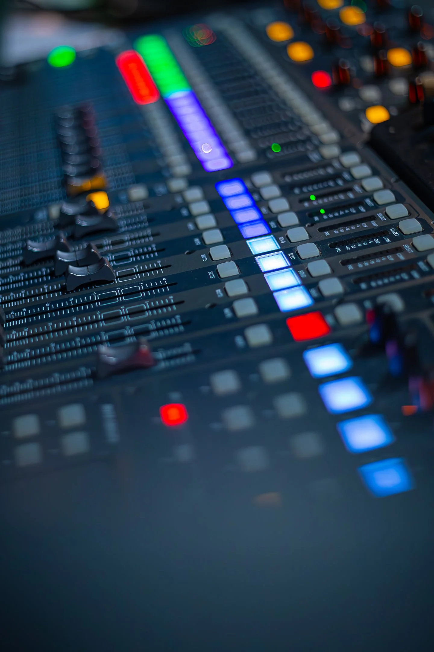 Close-up of a sound mixing board with illuminated faders and buttons in blue, green, red, and yellow, used for audio control.