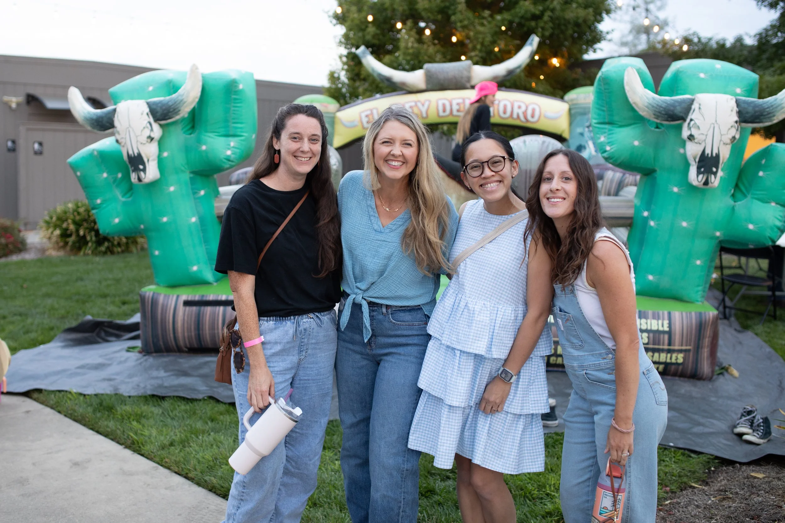 Four women smiling at the camera, standing outside in front of a carnival or fair game booth with inflatable bull sculptures and a sign that says "El Toro" in the background.