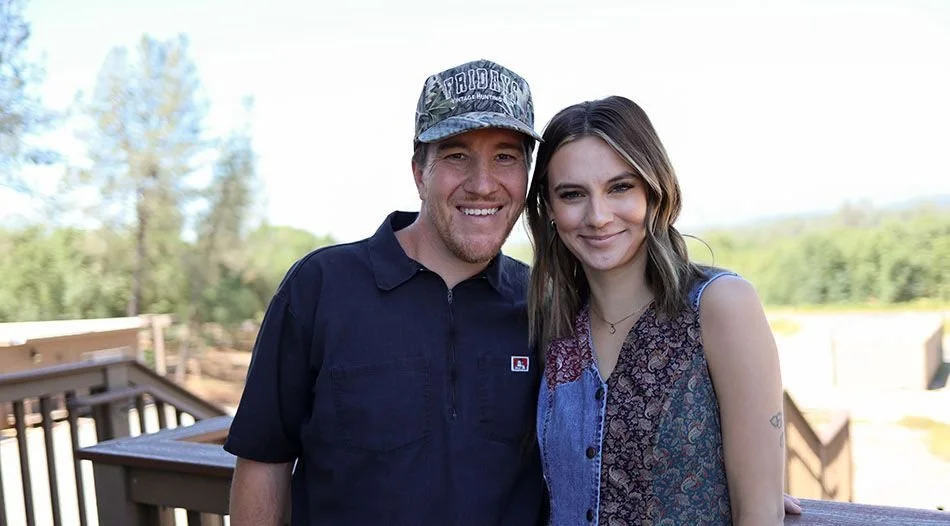 A man and a woman standing close together outdoors, smiling at the camera, with trees and a wooden deck in the background.