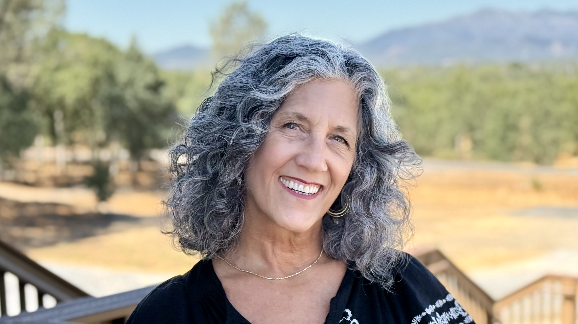 A smiling woman with curly gray hair outdoors, wearing a black top and gold jewelry, with a background of trees and mountains.
