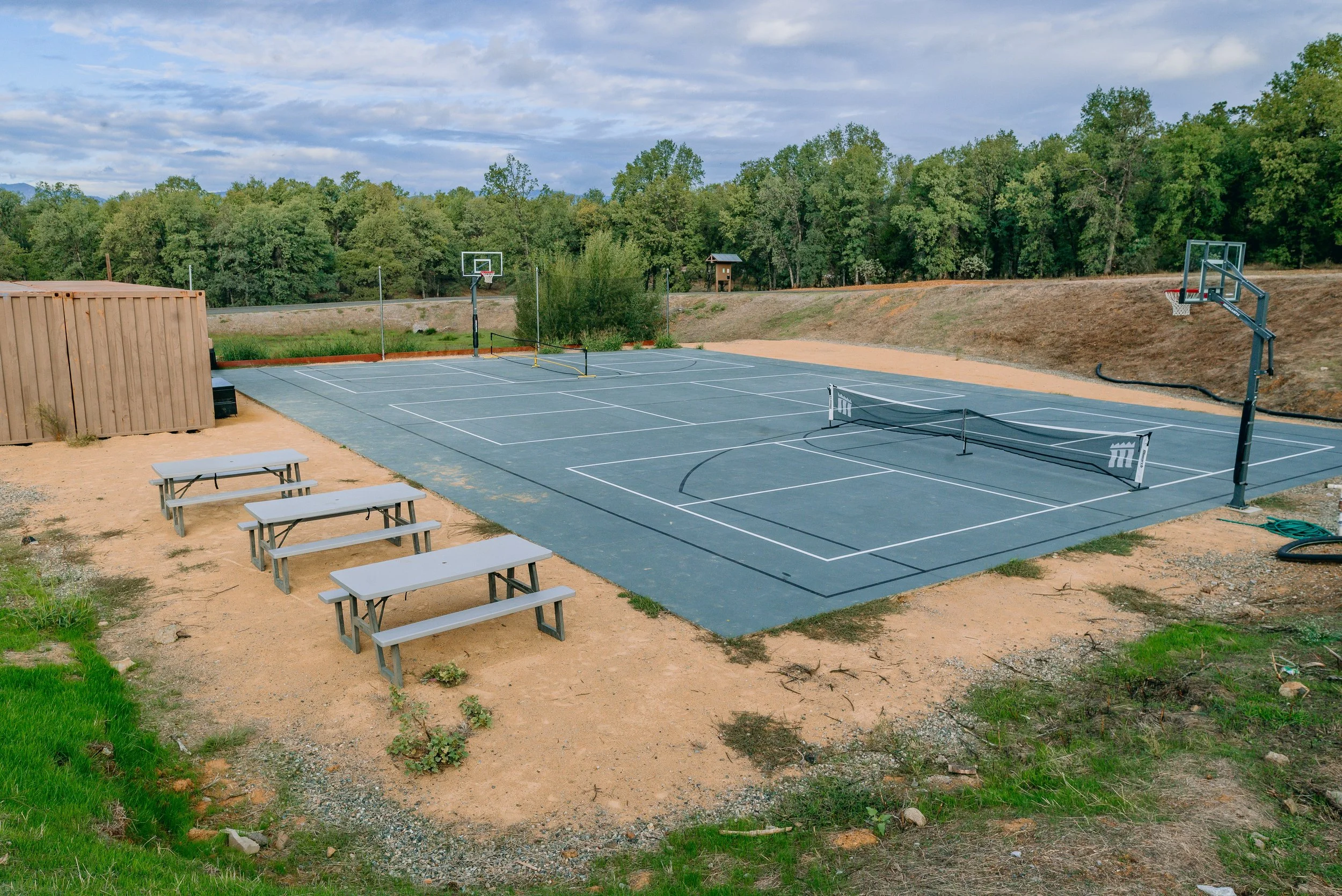 An outdoor basketball court with a net, hoops, and a divider, surrounded by trees and a trail, under a partly cloudy sky.