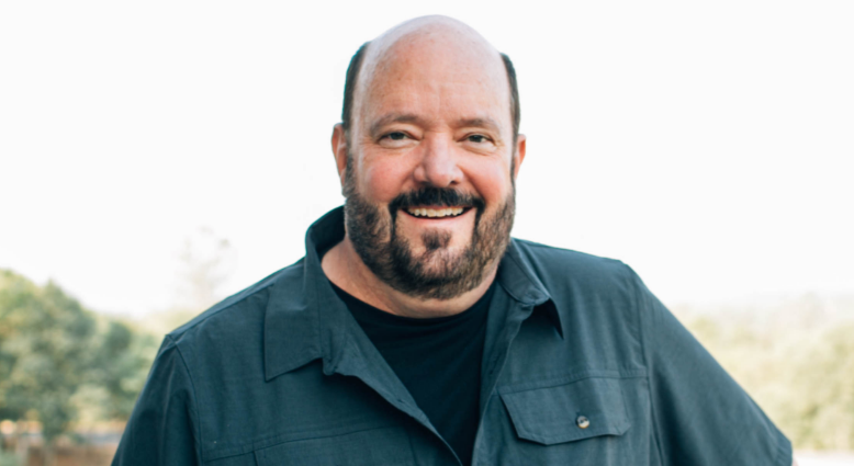 A smiling man with a beard and short hair, wearing a dark jacket, standing outdoors with a blurred natural background.