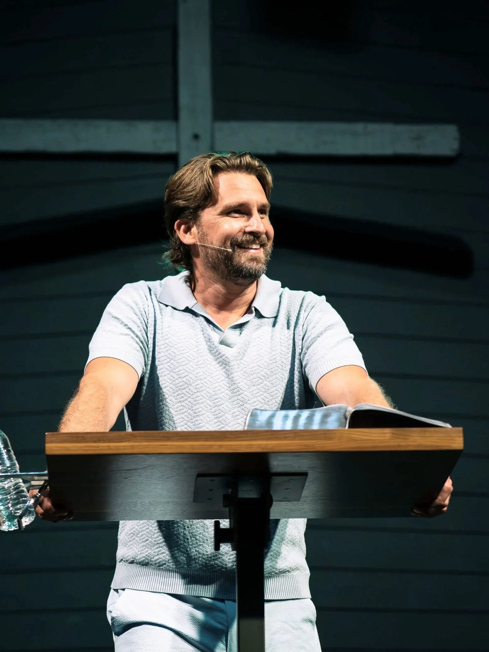 A man with brown hair, beard, and mustache smiling while sitting at a table with an open book, wearing a grey polo shirt, in front of a dark background with horizontal wooden panels.