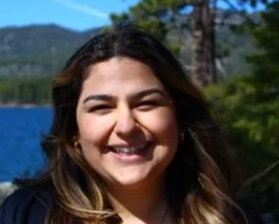Smiling woman outdoors in front of a lake and trees, with mountains in the distance.
