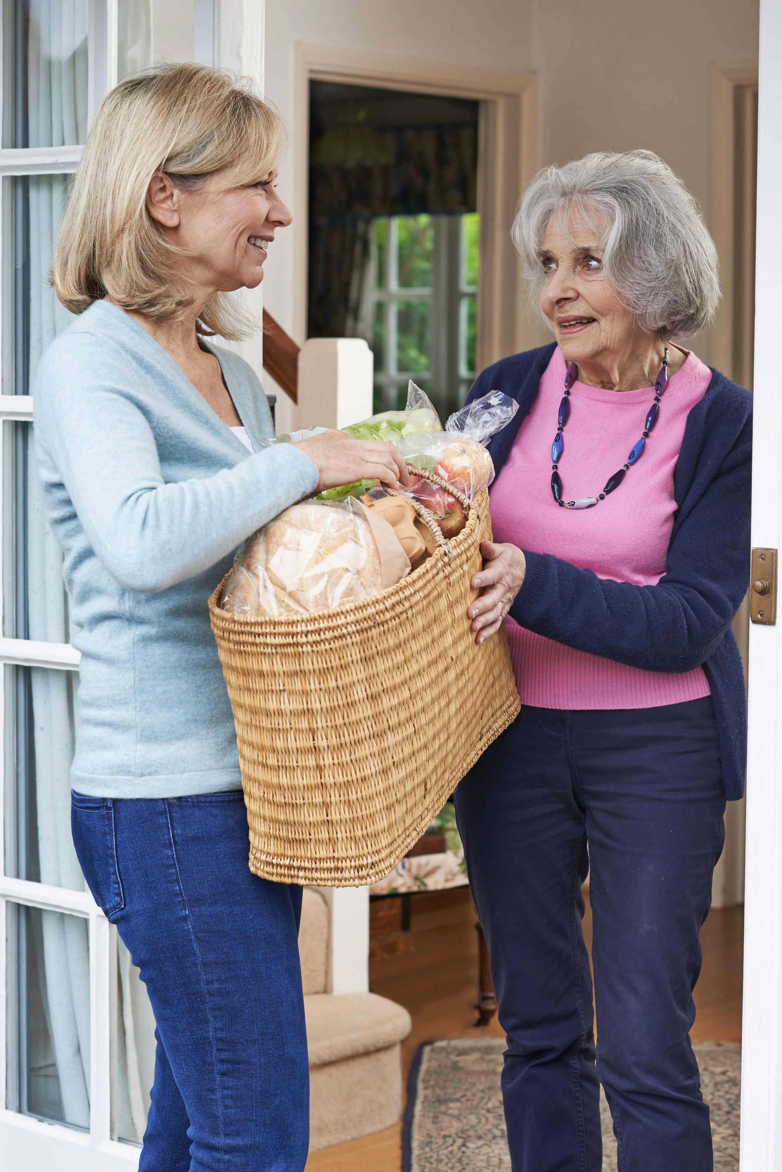 Two women exchanging a grocery bag at the front door of a house, smiling and engaging in conversation.