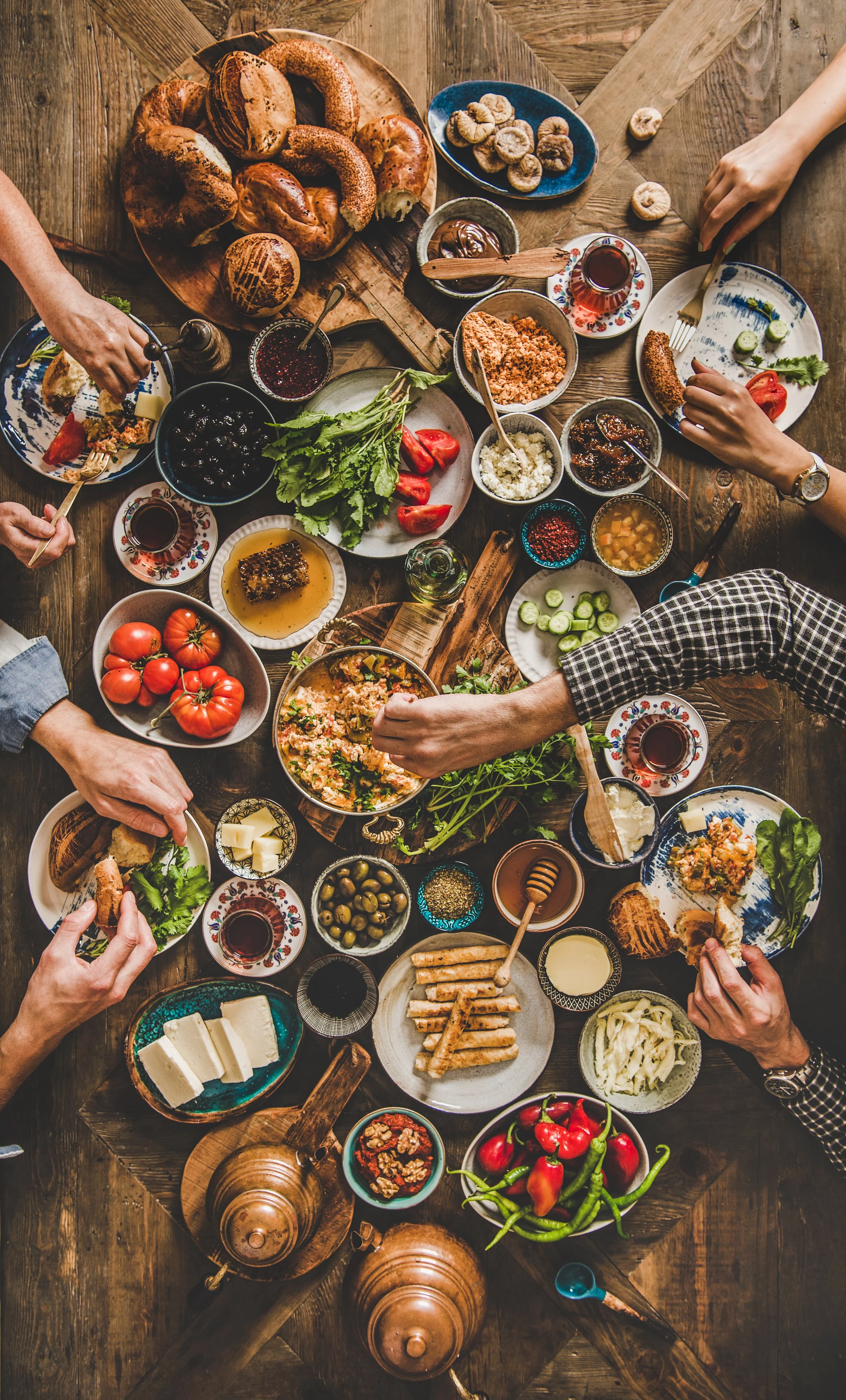 A top-down view of a wooden table filled with various dishes, bread, vegetables, cheeses, spreads, and beverages, with people sharing a meal.