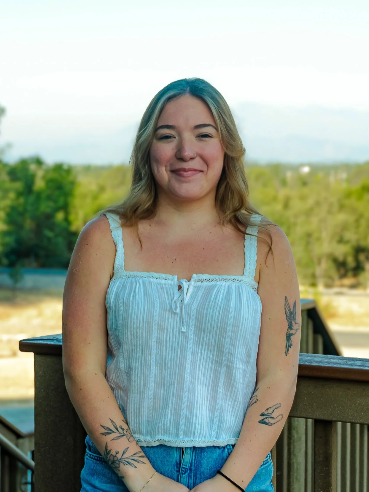 Smiling woman with tattoos on her arms, wearing a white sleeveless top, standing outdoors on a balcony with trees and a blue sky in the background.