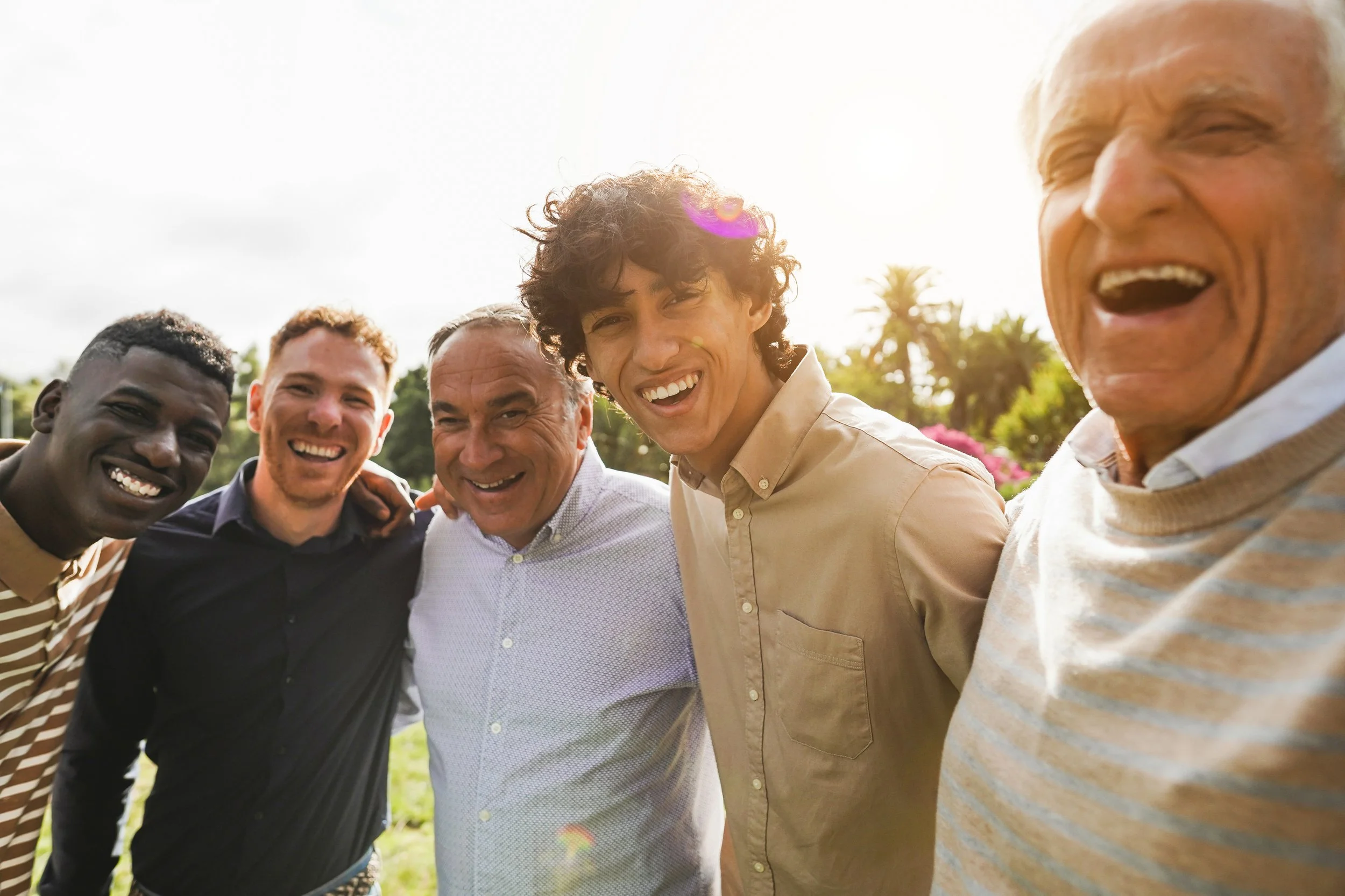 A group of five men standing close together outdoors, smiling and enjoying themselves. They are dressed casually, and the background features green trees and a bright sky with the sun shining.