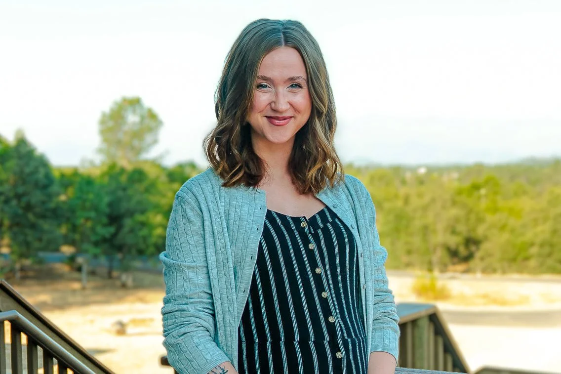 A woman with shoulder-length wavy brown hair and light skin, wearing a striped black and white top with gold buttons and a blue cardigan, standing outdoors on a balcony with green trees and blue sky in the background, smiling at the camera.