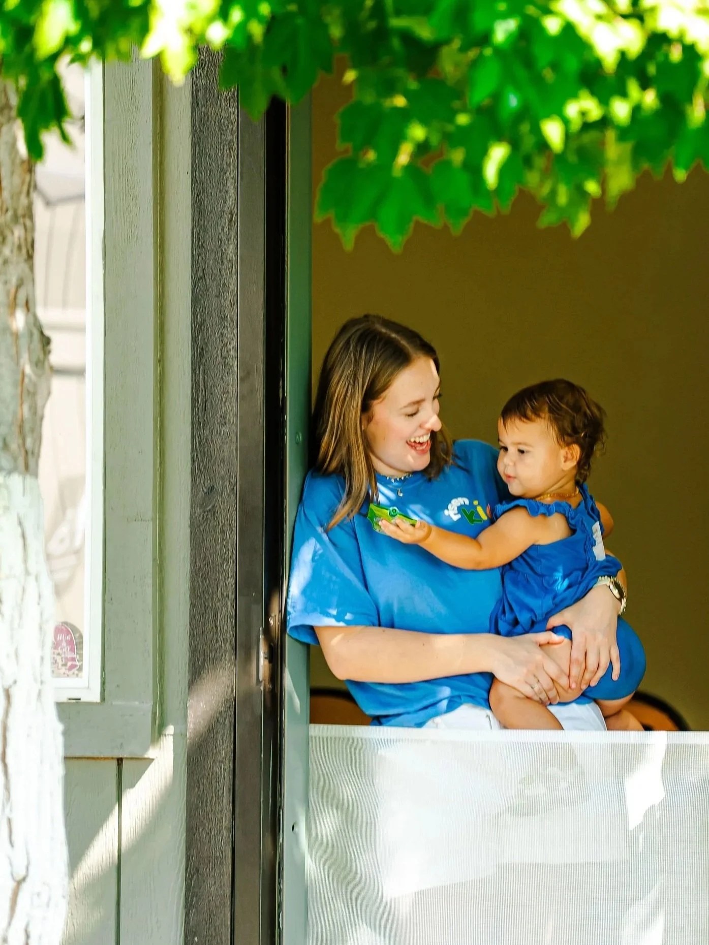 A woman holding a young child, both wearing blue shirts, smiling and looking at each other near a building with a light-colored wall and green foliage overhead.