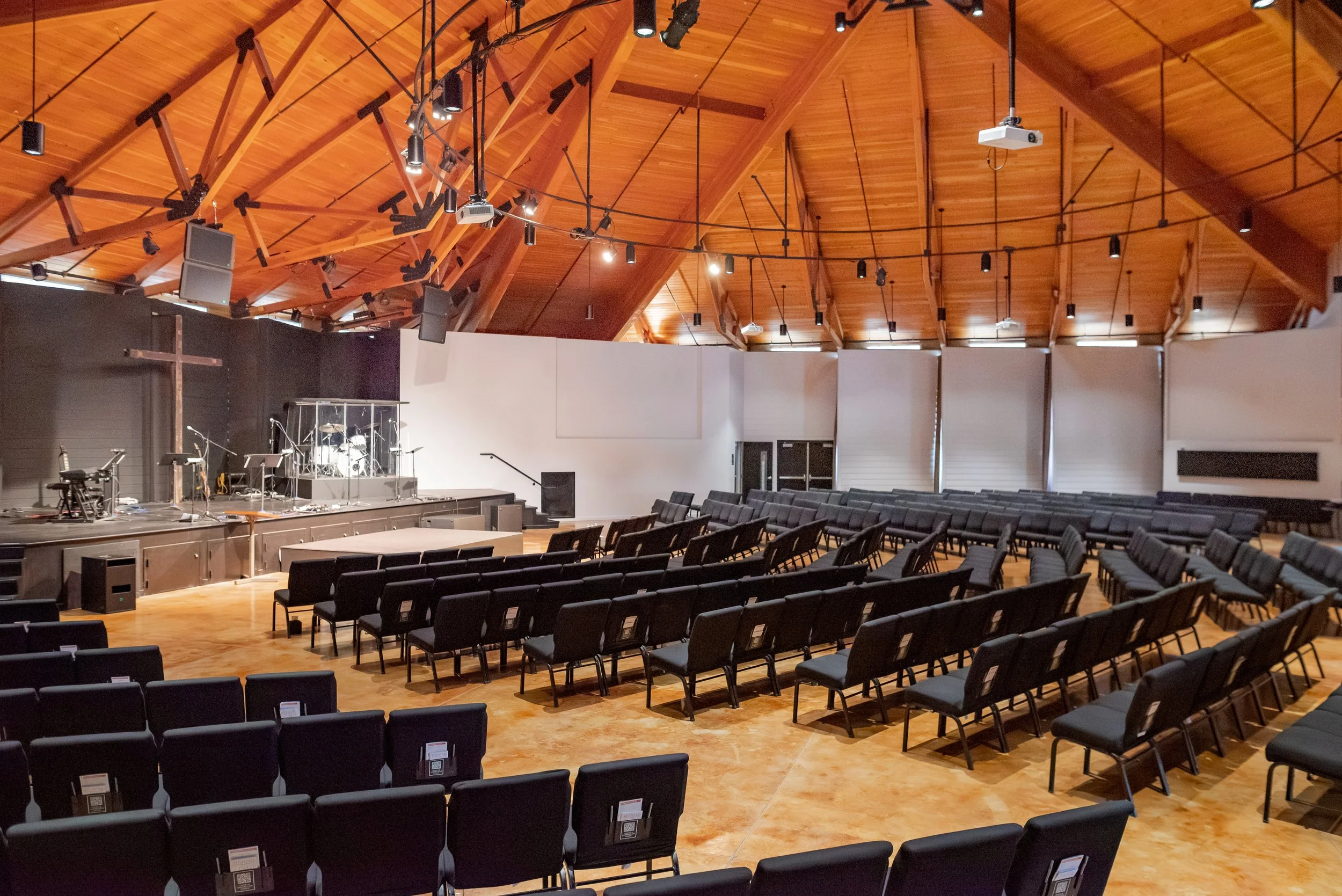 Interior of a church or auditorium with rows of black chairs, a stage with musical instruments, and a wooden ceiling with hanging lights and speakers.