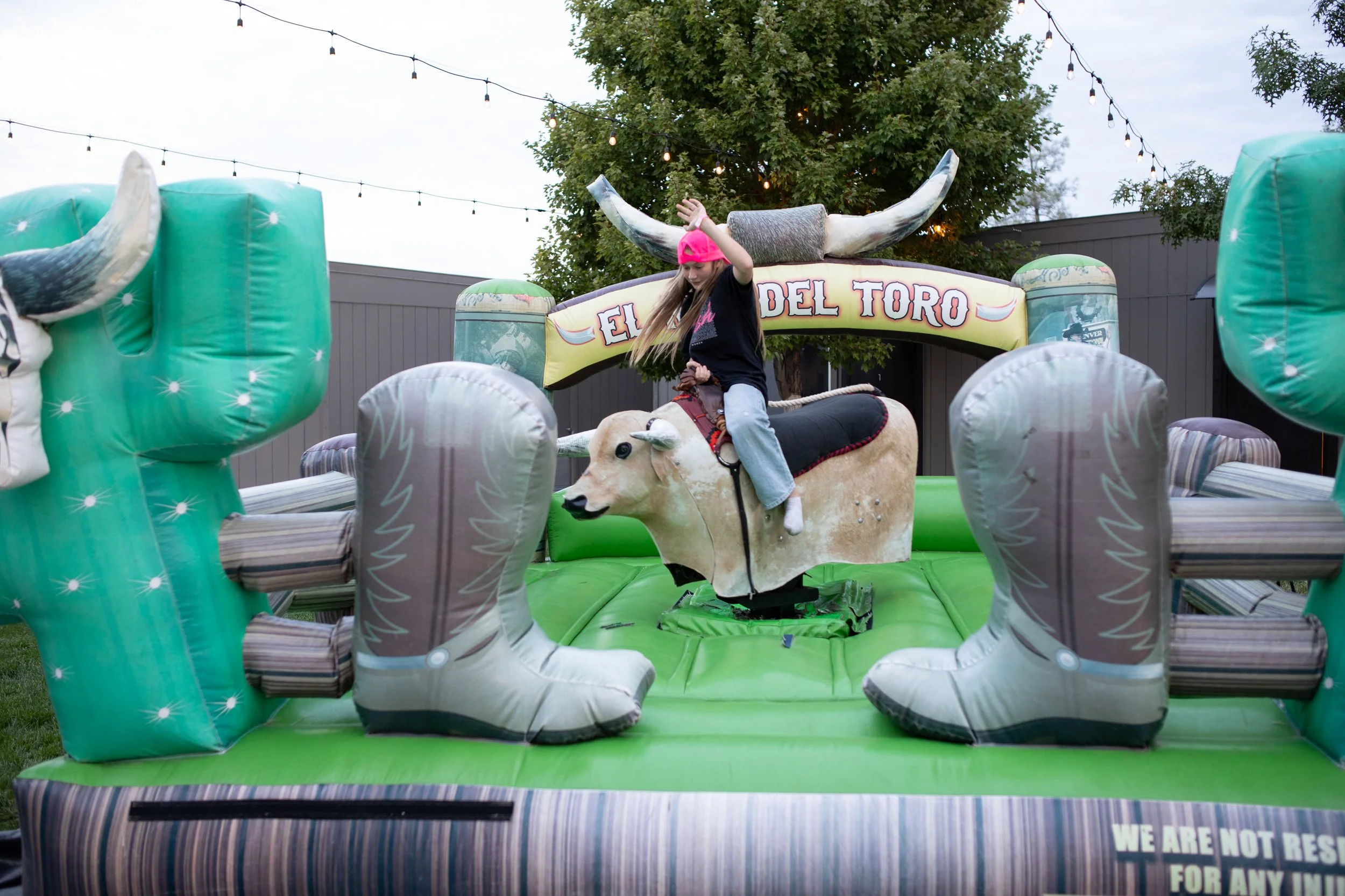 A girl wearing a pink cap riding a mechanical bull at an outdoor event. The setup includes inflatable cacti and a sign that reads "El Toro" with string lights hanging overhead.