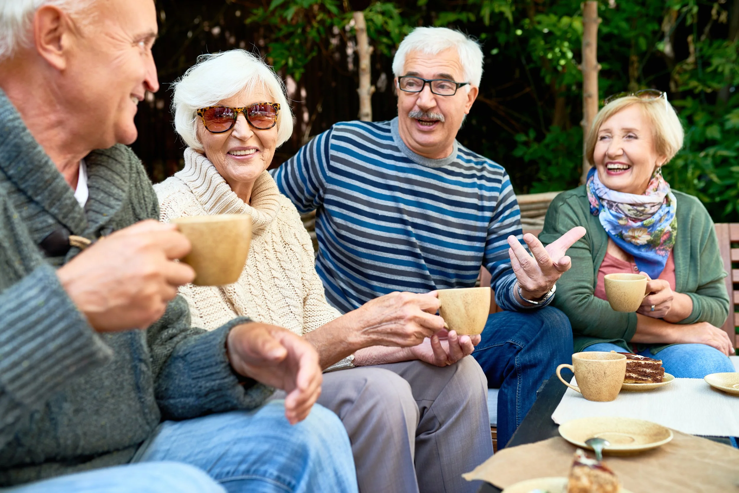 Group of elderly people sitting outdoors, chatting and laughing, with cups and plates of cake on the table in front of them.
