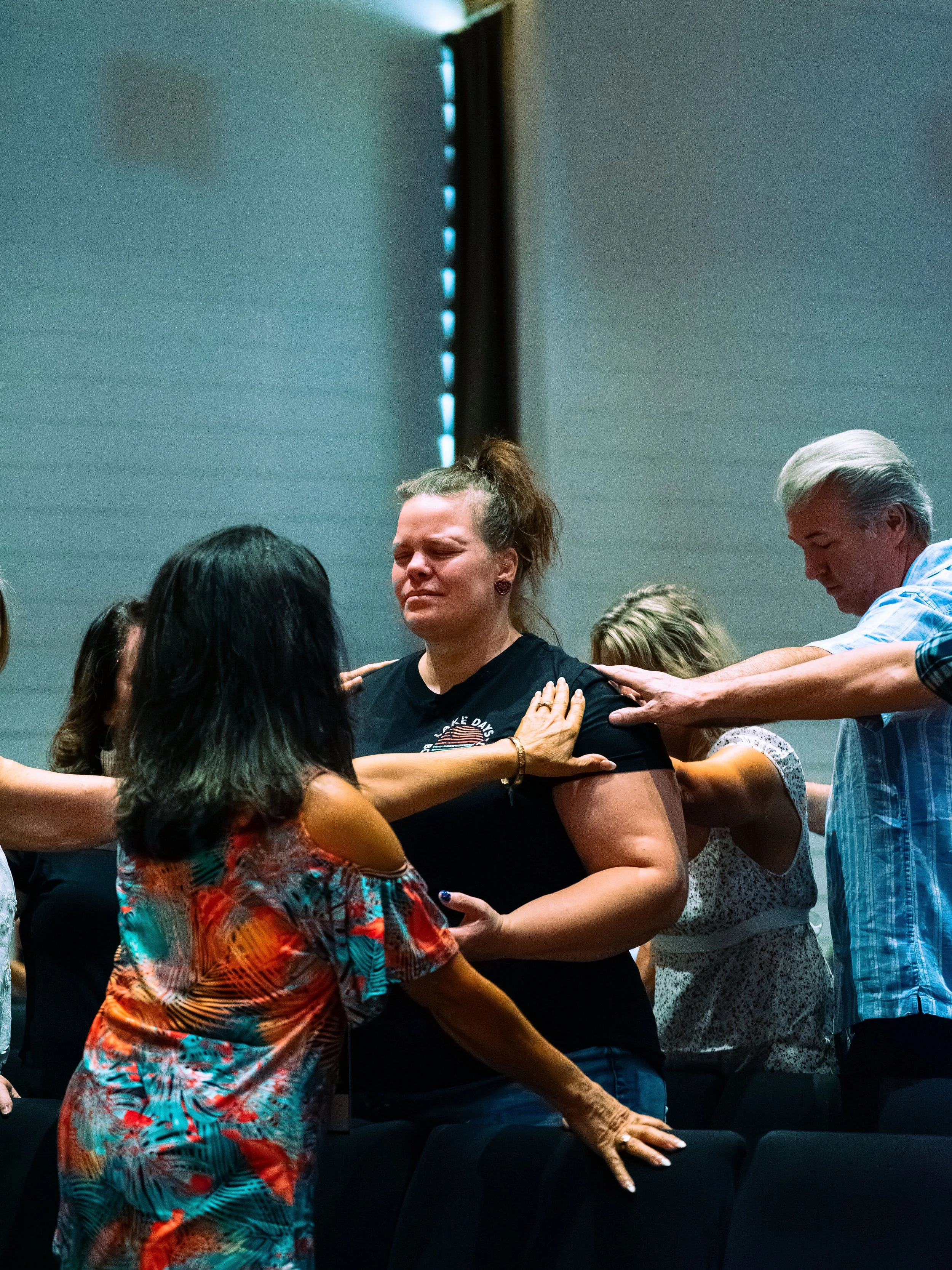 A group of people, mainly women, engaged in prayer or spiritual activity with their hands on each other's shoulders or arms, with a woman with a ponytail at the center, eyes closed in prayer, in a room with a gray wall and a black curtain.