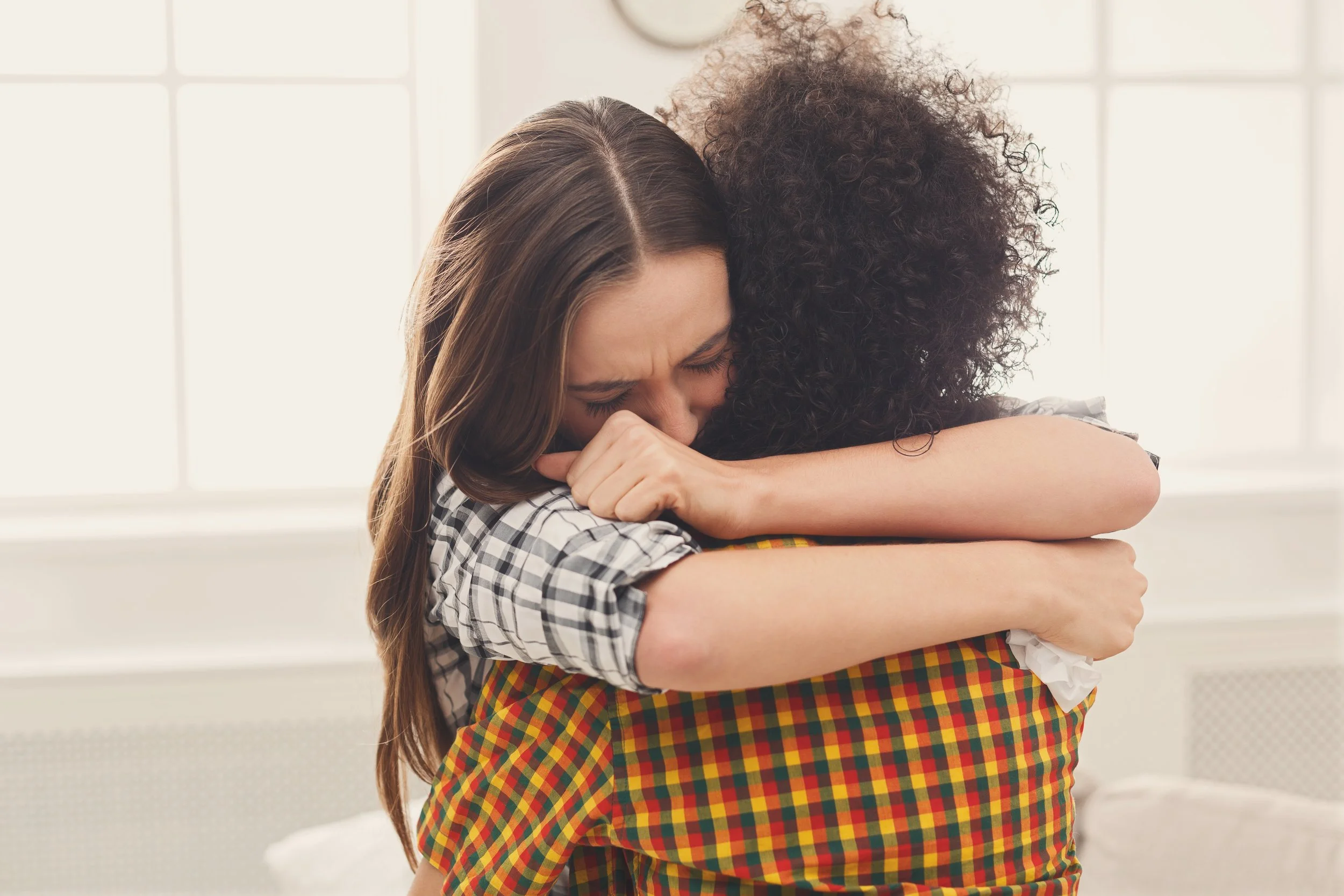 Two young women hugging each other, one with long brown hair and the other with curly dark hair, both appearing emotional in a bright room with large windows.