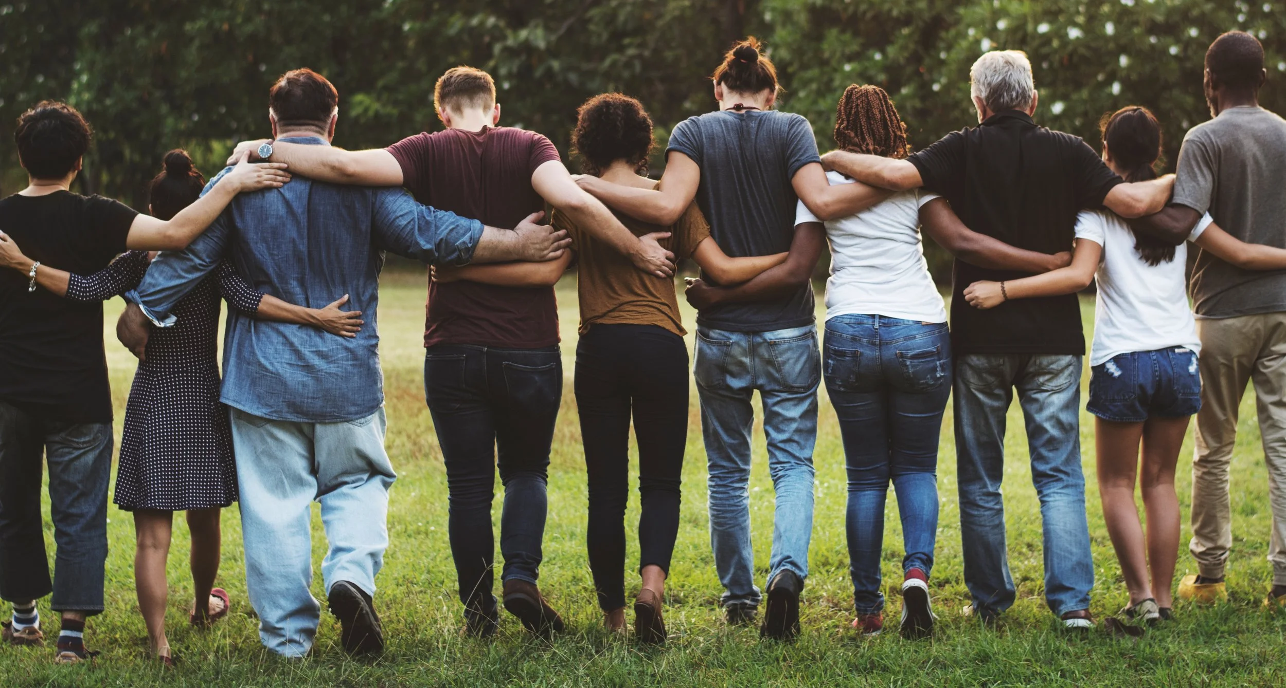 A diverse group of people holding each other by the shoulders and walking together outdoors on a grassy field during daytime.