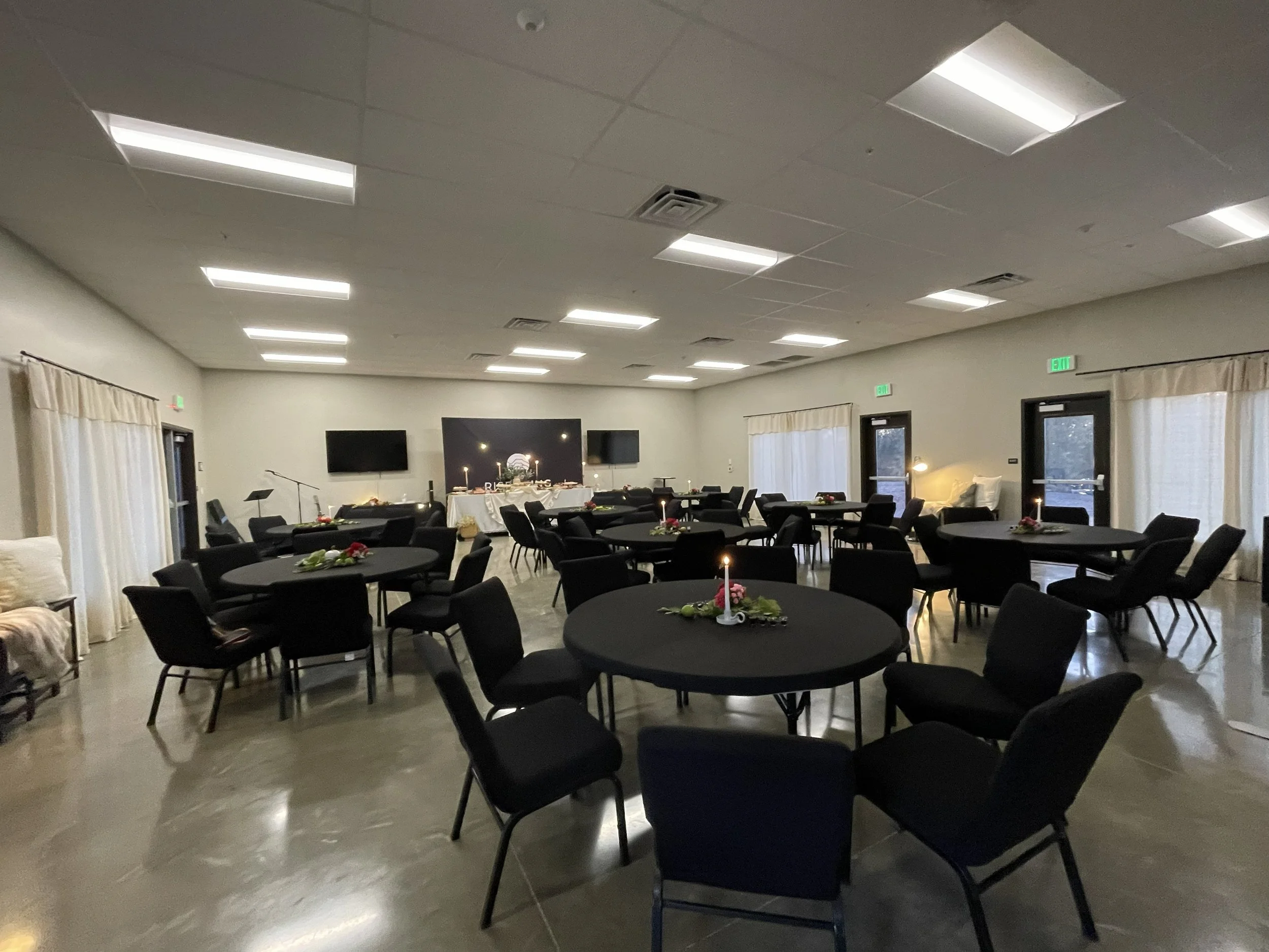 A banquet or event room with round tables covered in black tablecloths, each decorated with a small floral centerpiece and a candle. The room has a high ceiling with rectangular fluorescent lights, light-colored walls with draped curtains, and multiple exit doors with illuminated green exit signs.