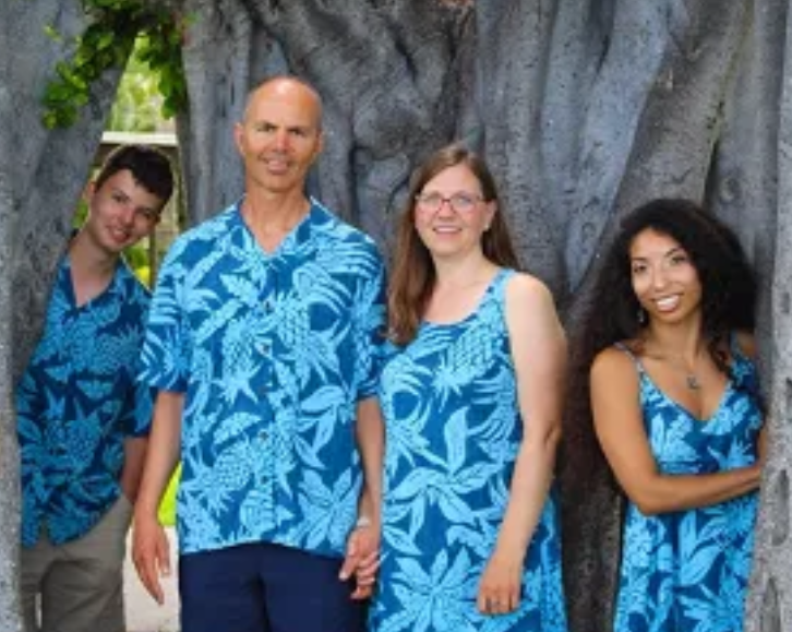 Four people standing outdoors in front of a large tree, wearing matching blue Hawaiian shirts or dresses with tropical leaf patterns.
