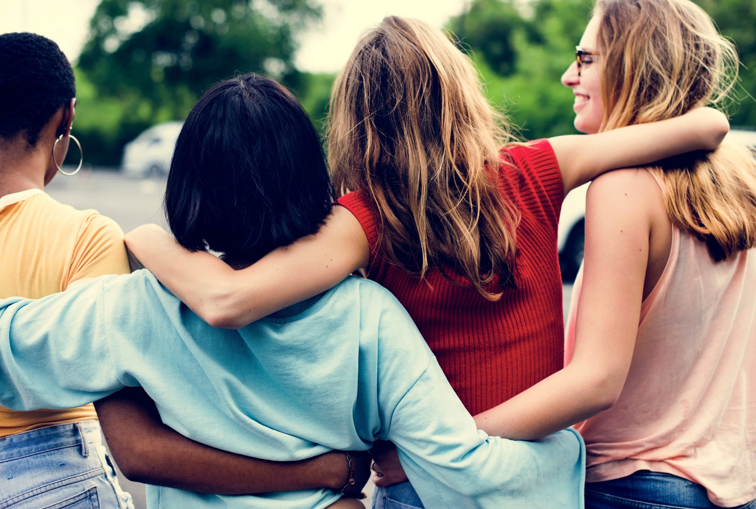 A group of four diverse women standing outdoors, hugging each other, and smiling.