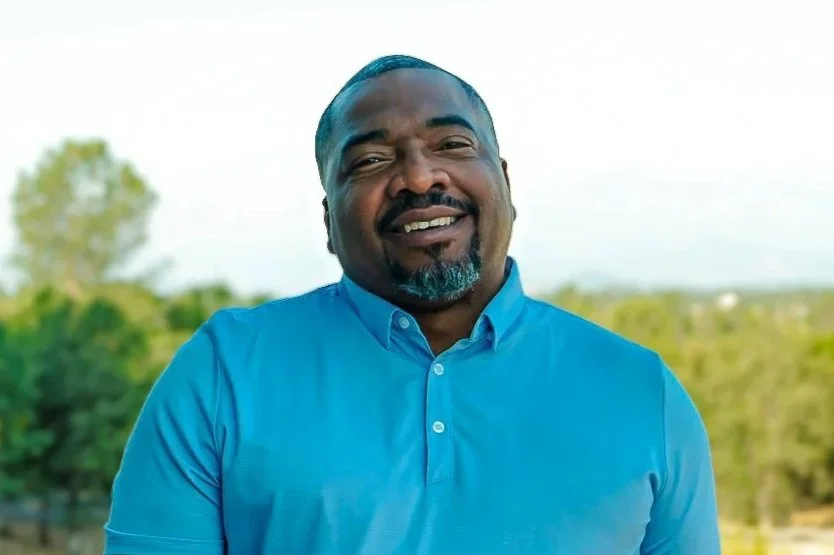 A smiling Black man with a goatee and mustache wearing a blue button-up shirt outdoors with trees in the background.