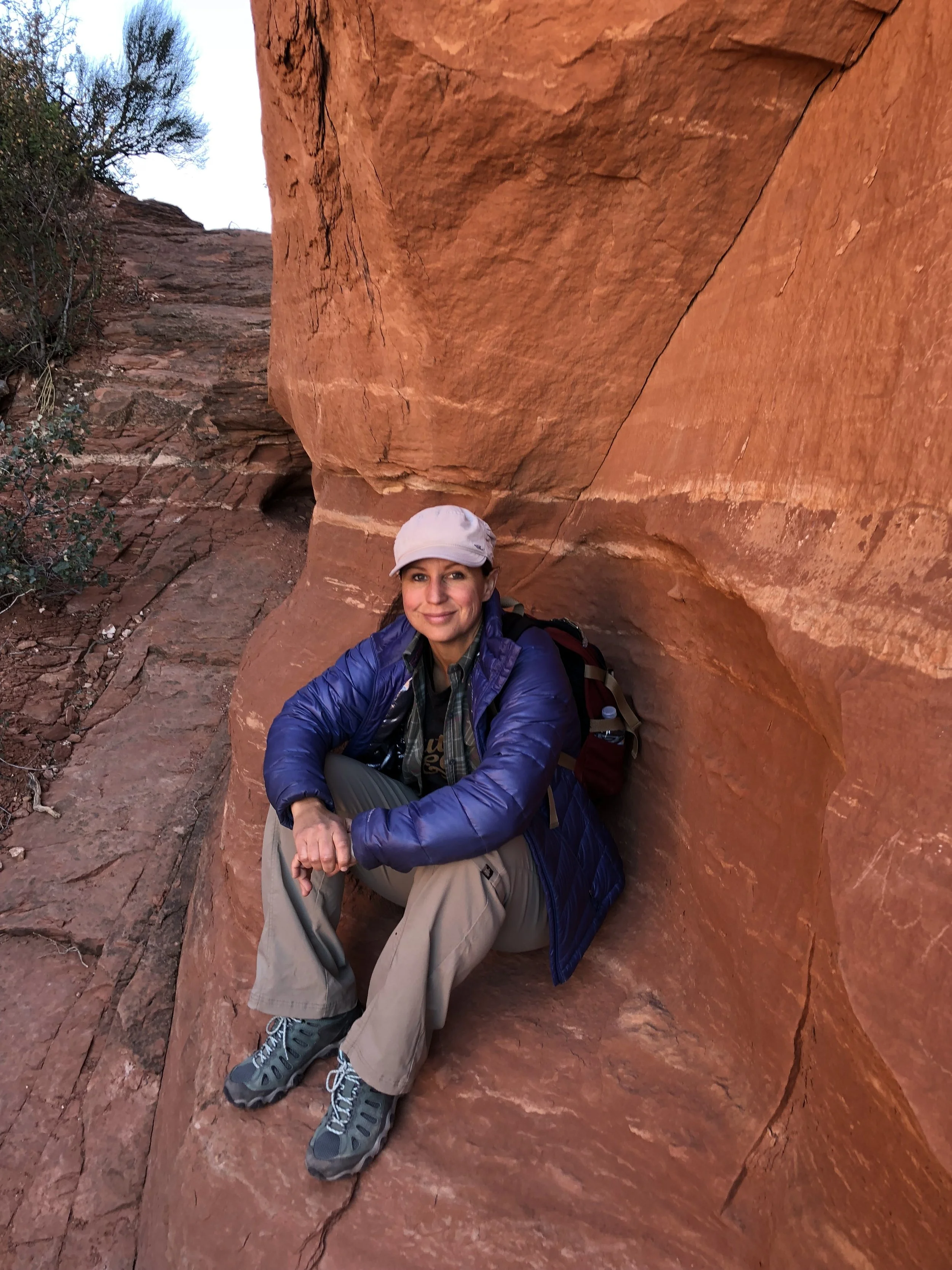 A woman sitting on the ground in front of large red rock formations during a hike.