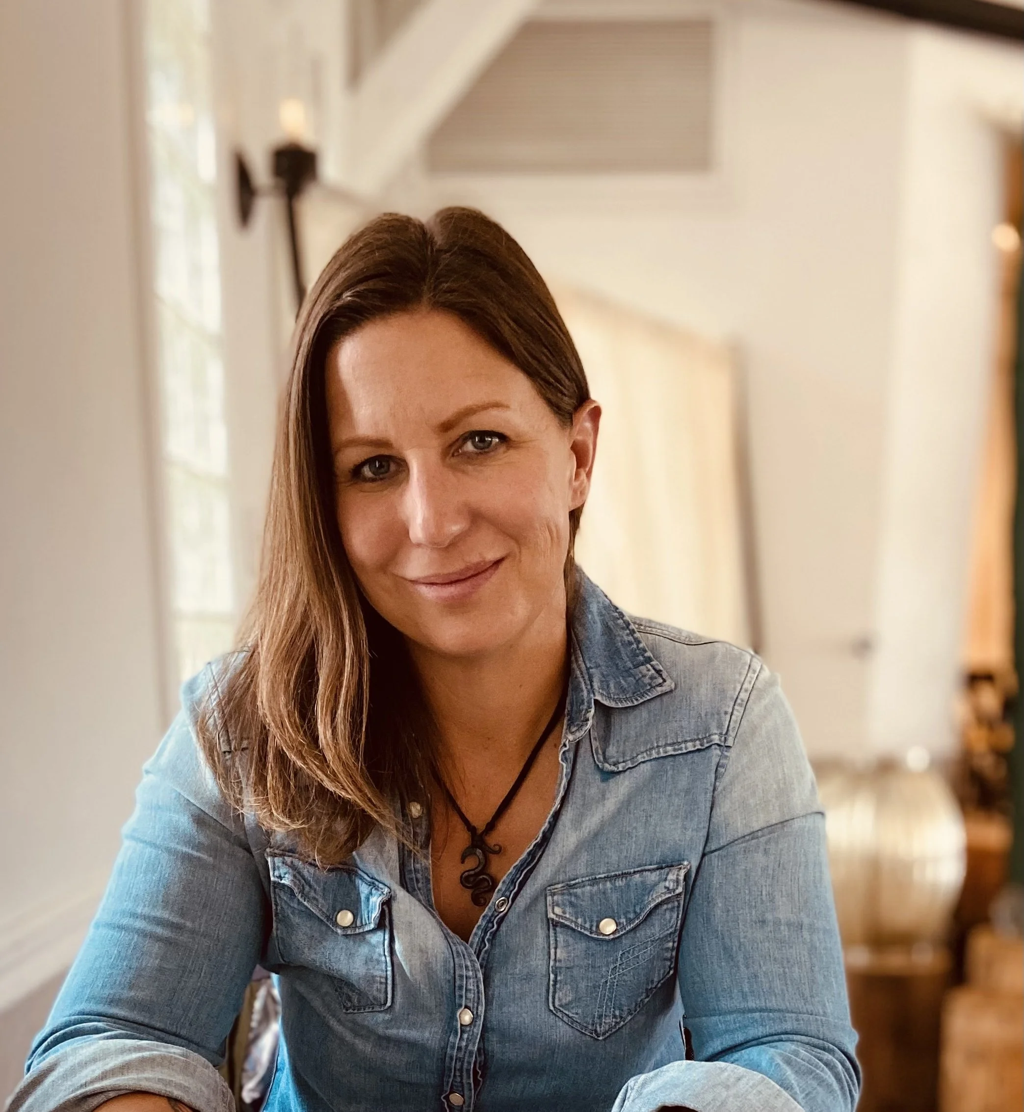 A woman with long brown hair and blue eyes wearing a denim shirt with rolled-up sleeves, sitting indoors with blurred background of a hallway and windows.