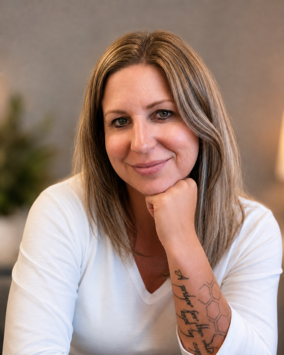 A woman with long, light brown hair and fair skin, smiling gently, resting her chin on her hand, wearing a white shirt, with tattoos on her arm against a blurred indoor background.