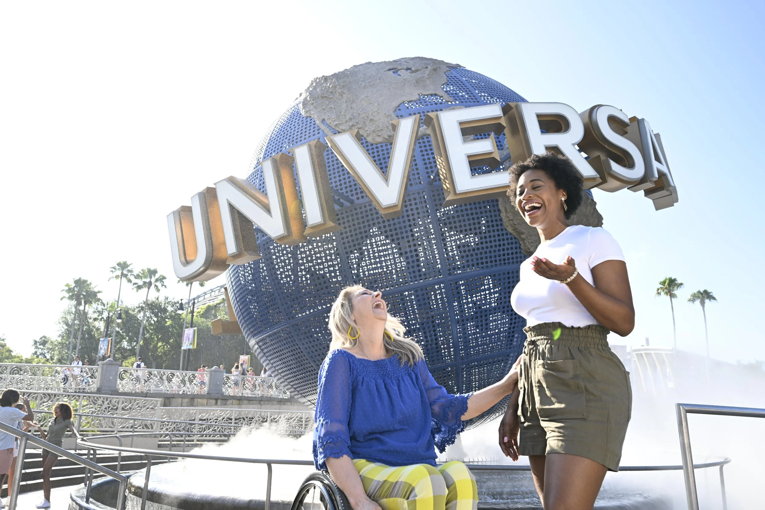 Two women, one sitting in a wheelchair and wearing a blue top and yellow striped pants, and another standing and wearing a white top and gray shorts, are posing and smiling in front of the Universal Studios globe.