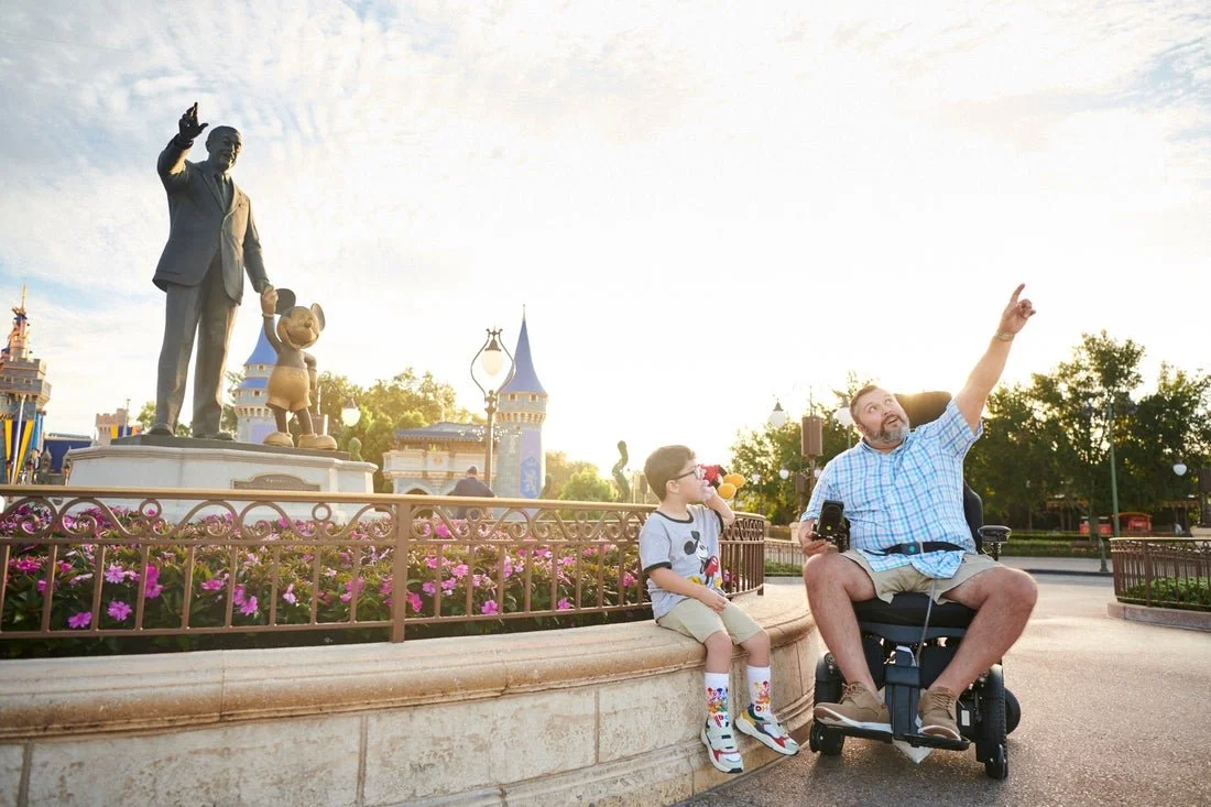 A man and a young boy sitting on a circular stone ledge in front of a Disneyland statue of Walt Disney and Mickey Mouse, with a castle in the background, all outdoors during daytime.