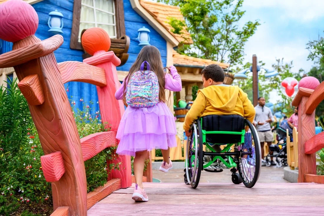 Two children, one girl in a purple dress with a backpack and one boy in a yellow hoodie in a wheelchair, walking on a wooden bridge in a colorful amusement park or playground with whimsical pink and brown fencing and a blue building in the background.