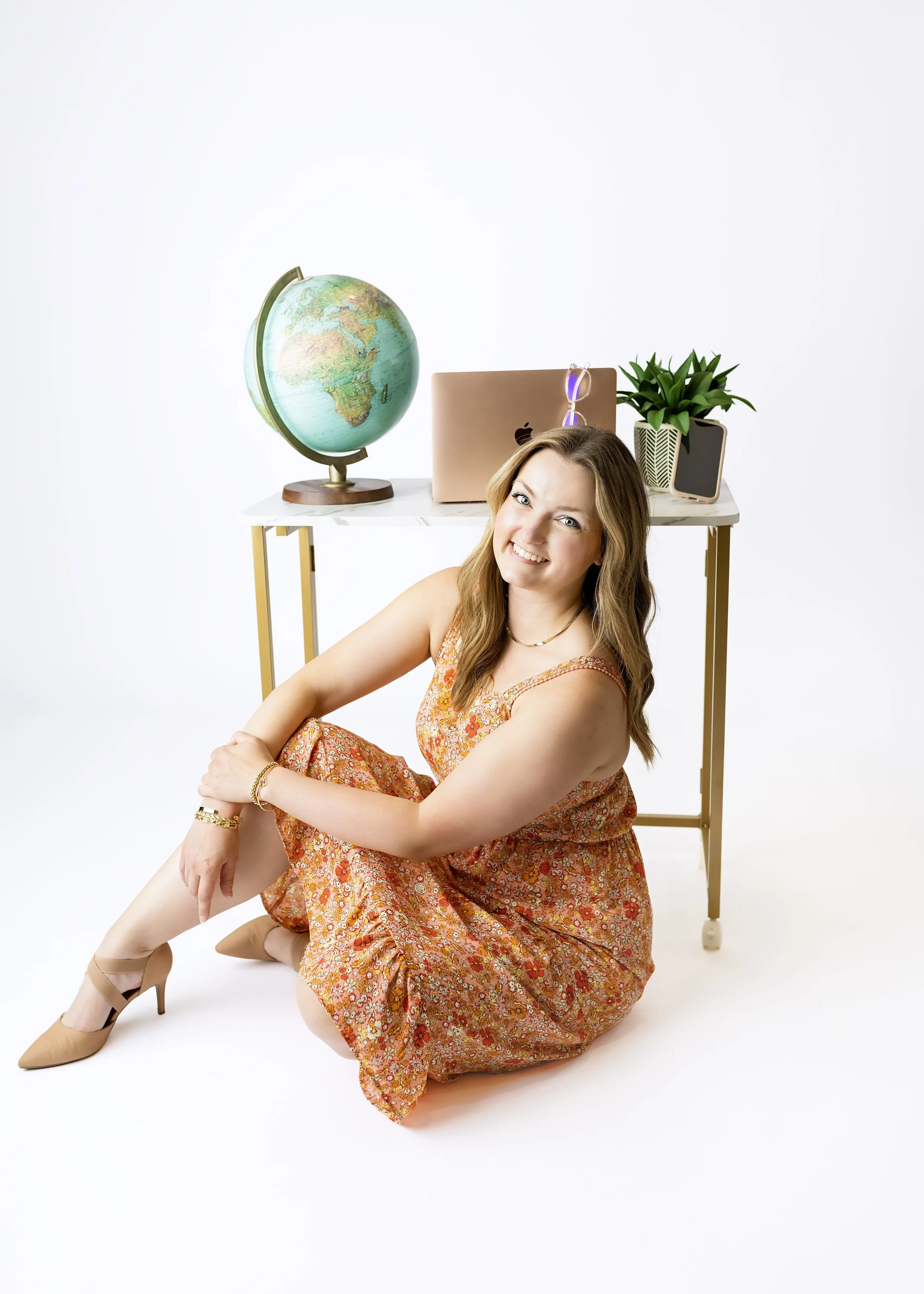 A young woman in an orange floral dress sitting on the floor with her legs crossed, smiling at the camera. Behind her, a white table holds a globe, a closed rose gold laptop with glasses on top, a small potted plant, and a smartphone.