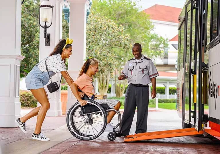 A young girl in a wheelchair being assisted onto a bus by two people, with a bus driver standing nearby.