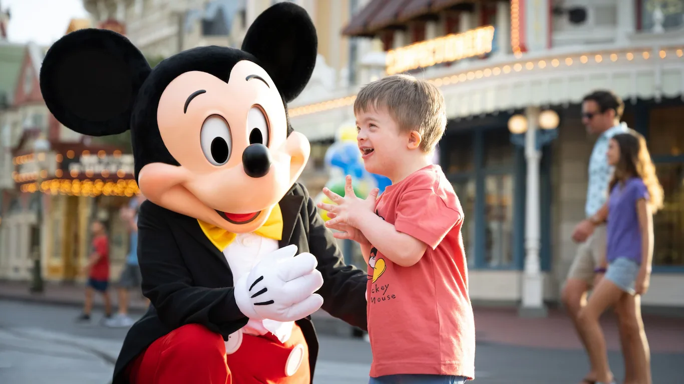A young boy in a red t-shirt is happily interacting with Mickey Mouse, who is dressed in a tuxedo with a yellow bow tie, on a street with other people and a building with lights in the background.