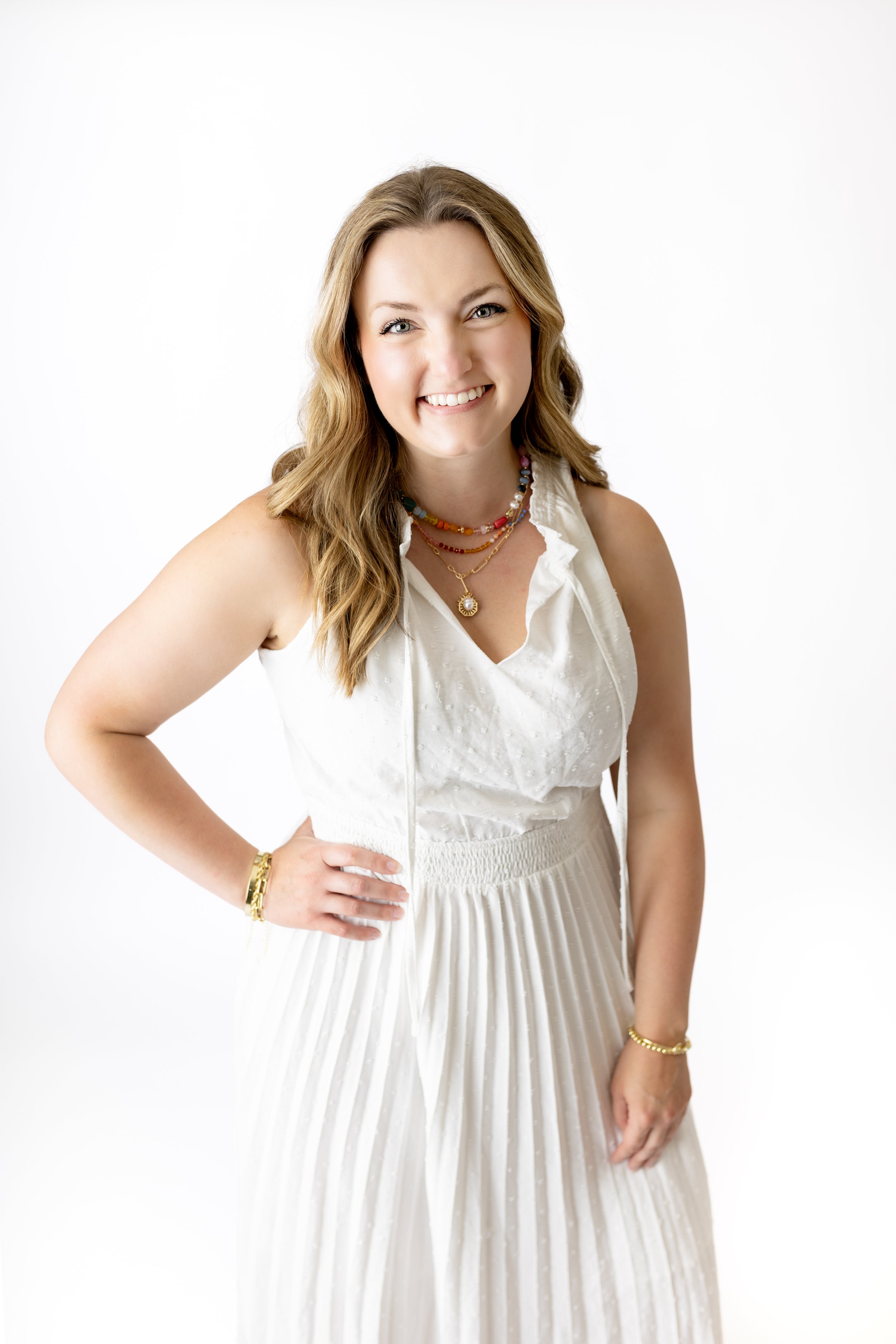 A woman with light skin, wavy light brown hair, and green eyes smiling, wearing a sleeveless white dress with pleats, multiple colorful necklaces, and gold bracelets, against a plain white background.
