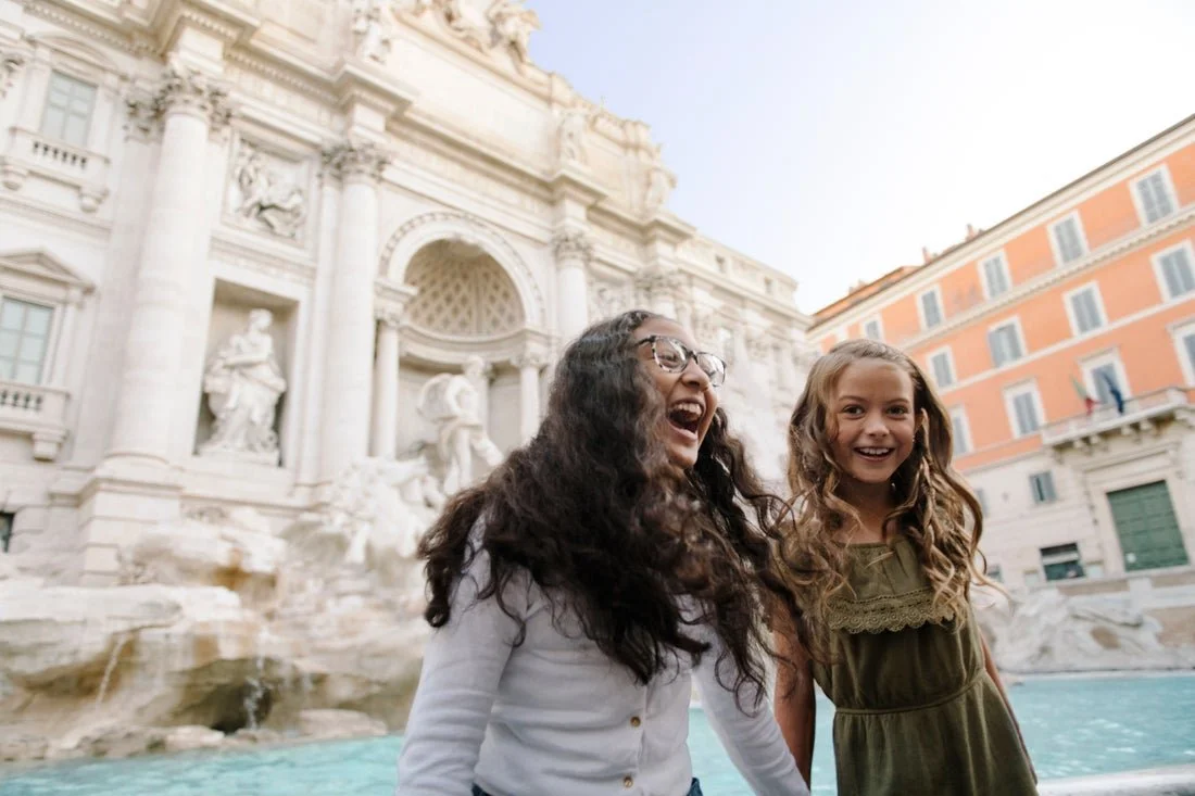 Two women smiling and laughing in front of the Trevi Fountain in Rome, Italy, during daytime.