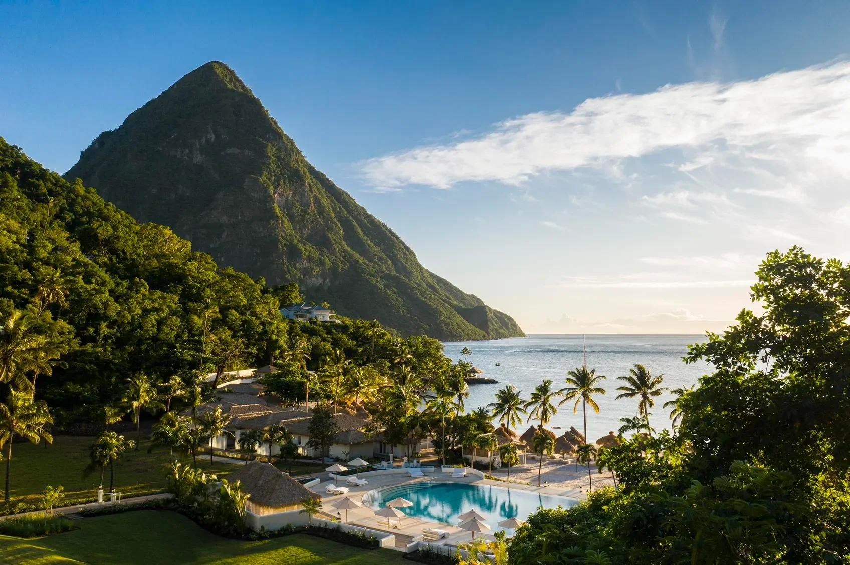 Tropical resort with a swimming pool, surrounded by palm trees and lush greenery, overlooking the ocean with a mountain in the background during sunset.