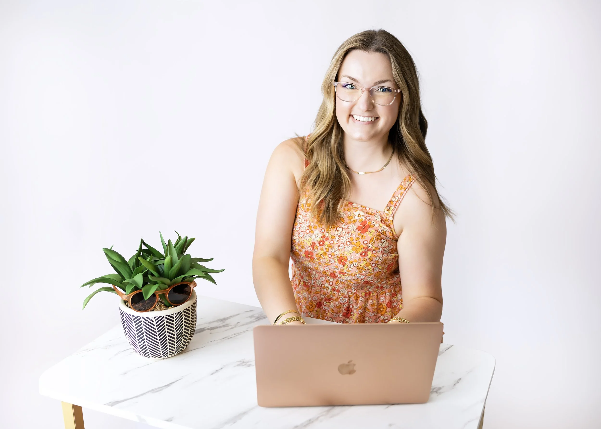 Woman with light brown hair and glasses sitting at a marble table with a pink MacBook, a potted plant, and sunglasses in a ceramic pot, smiling against a plain white background.