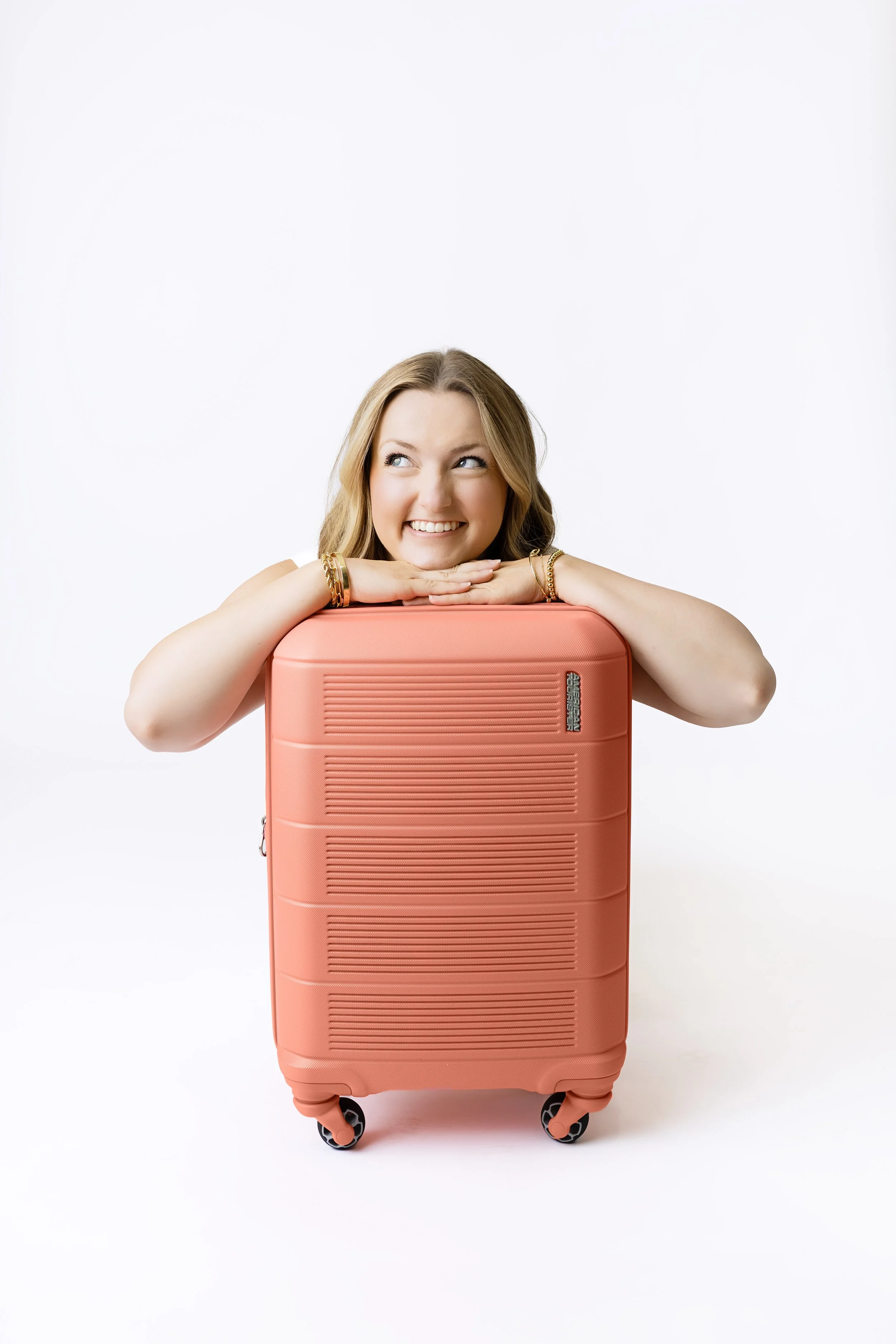 Young woman smiling and resting her chin on a pink rolling suitcase against a white background.