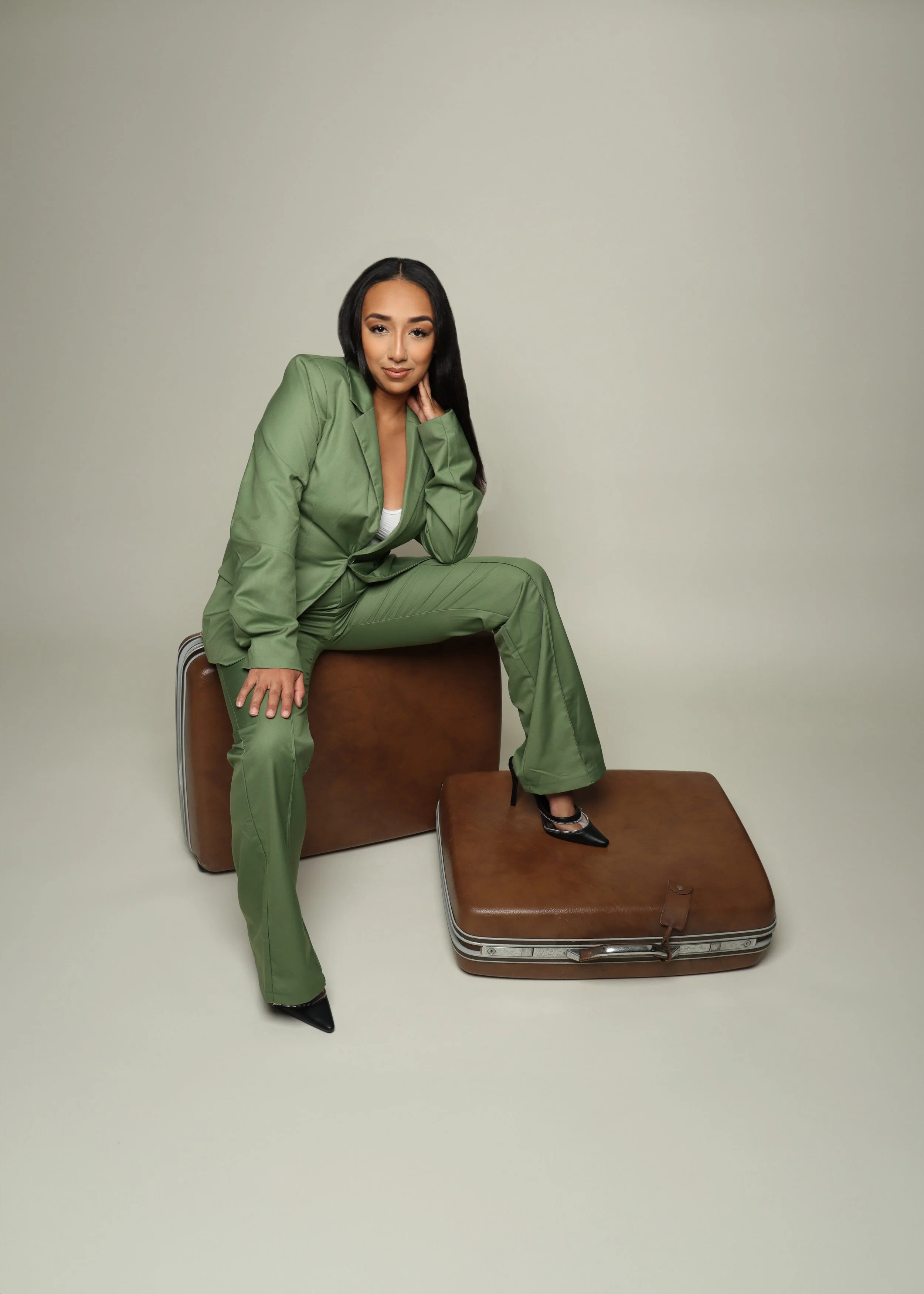 A woman in a green suit sitting on a suitcase and large lounge chair, with one leg propped on another chair, in front of a plain light-colored wall.