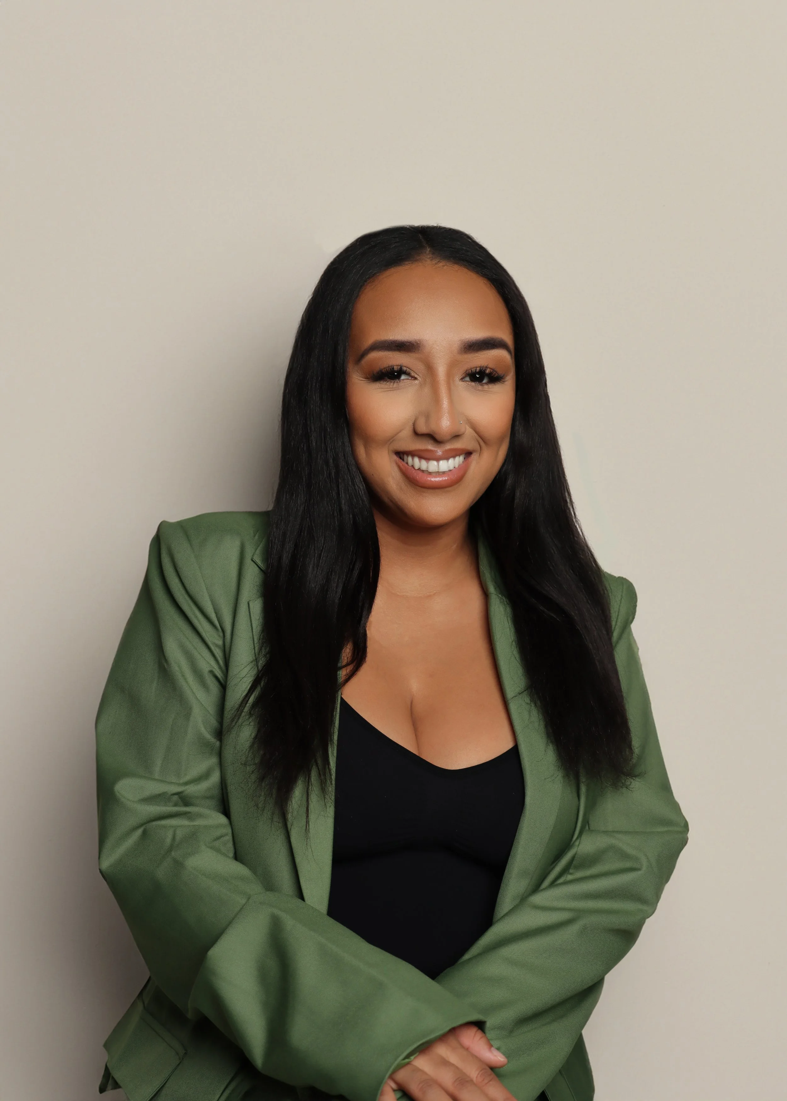 A woman with long black hair smiling, wearing a black top and a green blazer, standing against a plain white wall.