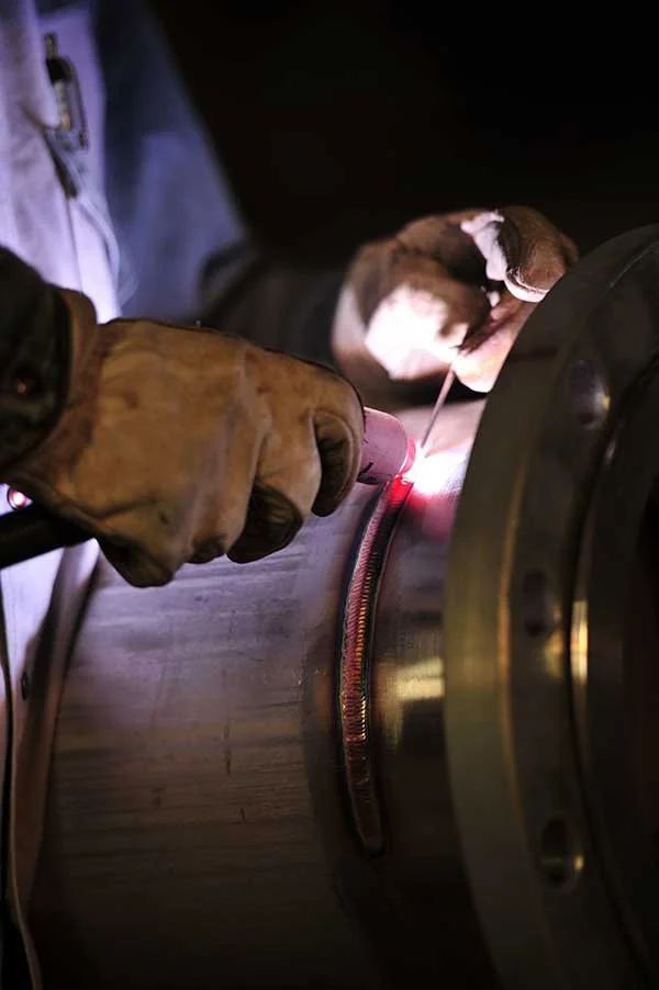 Welder wearing gloves and protective gear welding a metal pipe, with bright sparks and light from the welding torch.